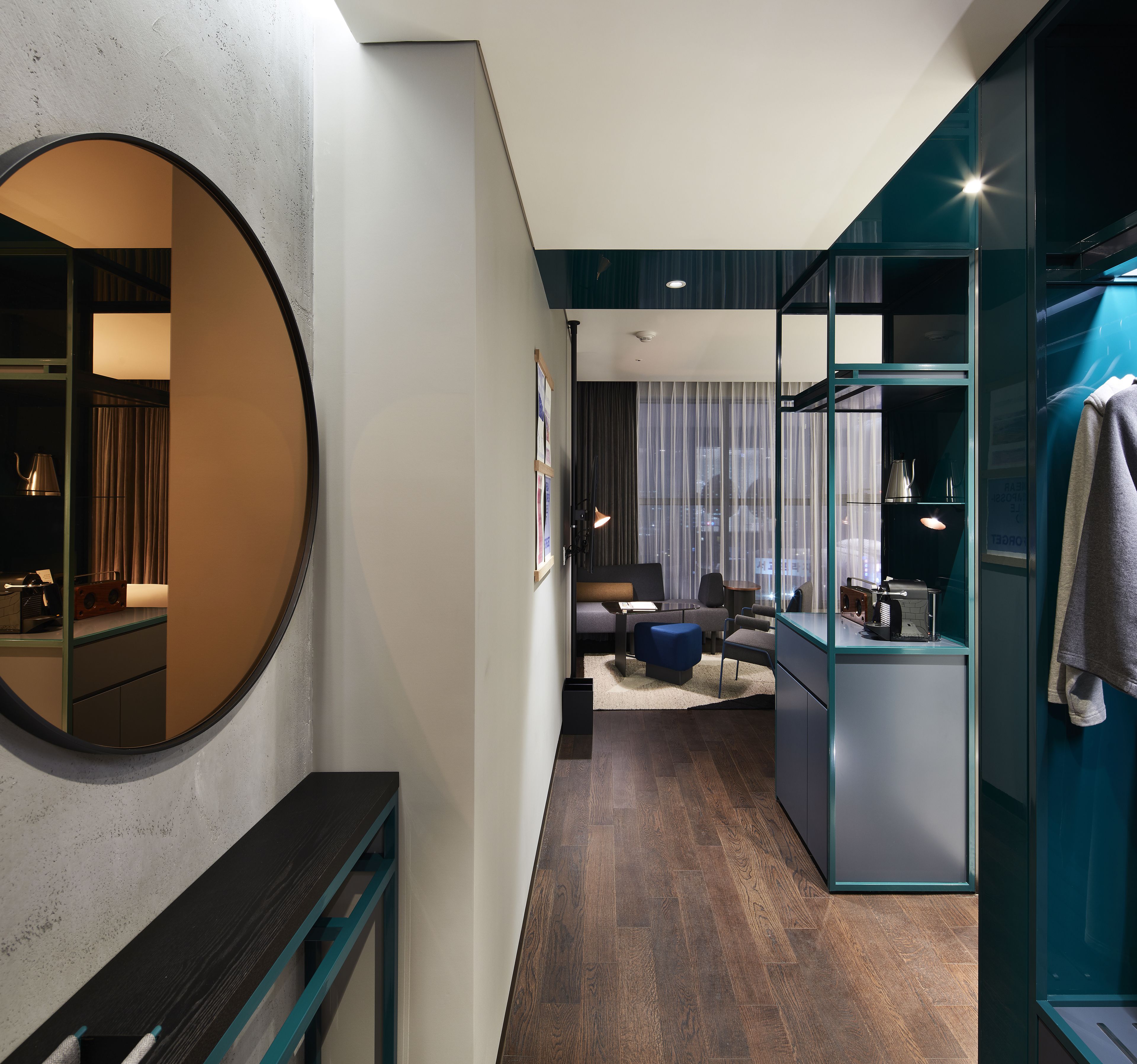 A modern hotel room with a round mirror, a dark wooden bench, a seating area with a sofa and ottoman, a kitchenette with a coffee machine, and a glass partition.