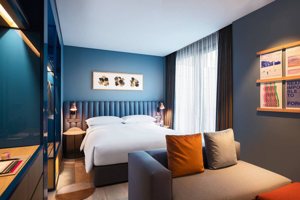 A modern hotel room with a large bed, a sofa, and a desk. There are artwork on the walls and a window with curtains.