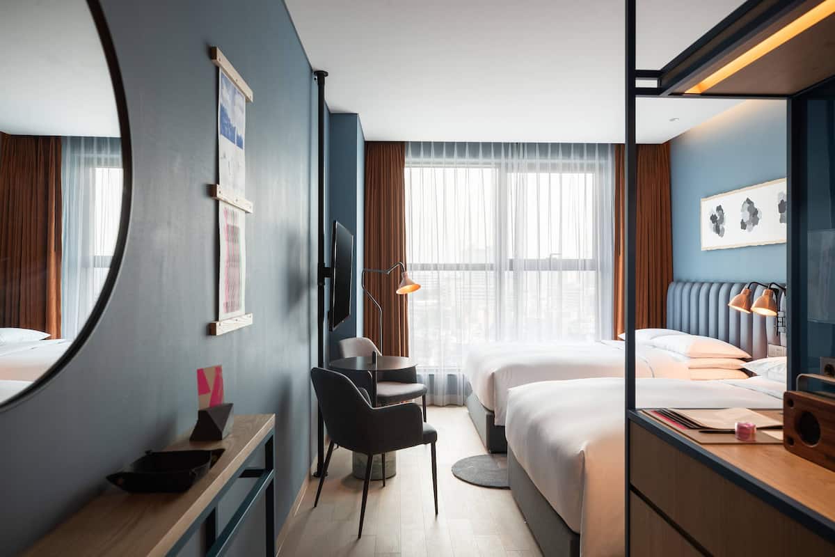A hotel room with two beds, a desk, a chair, a round mirror, and a large window with curtains.