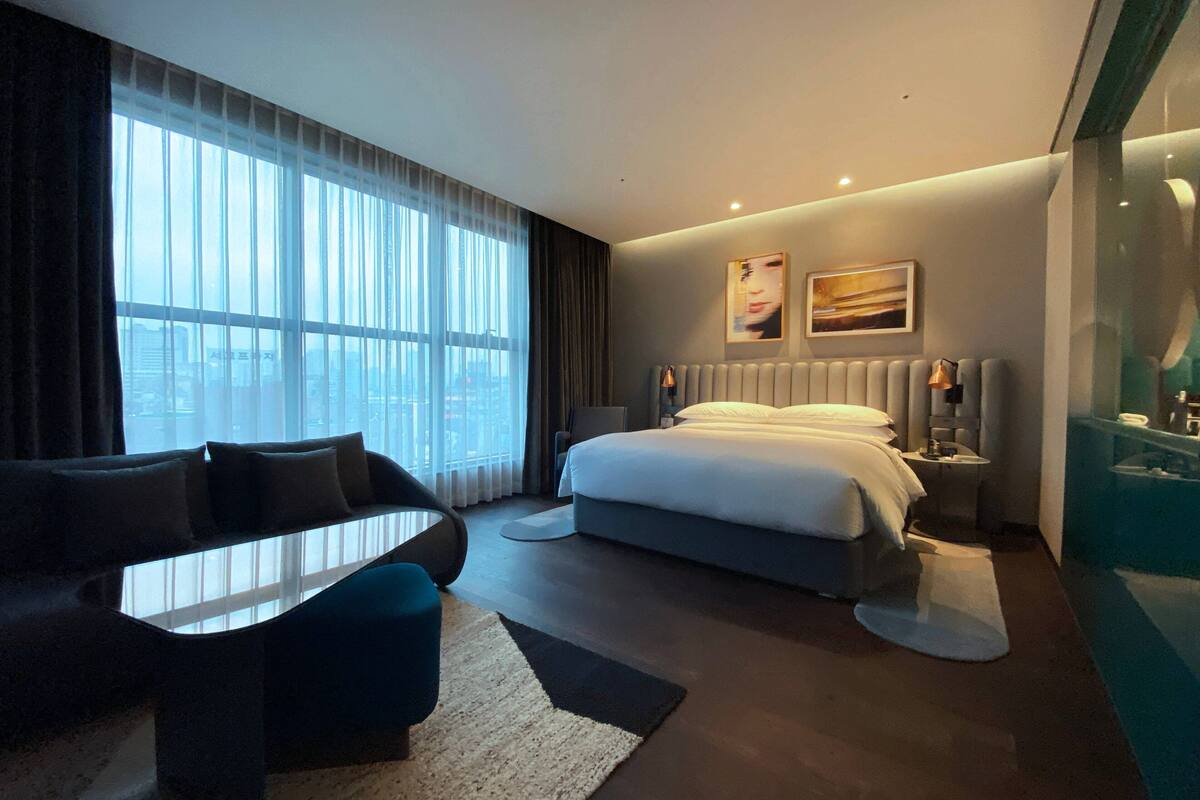 A modern hotel room with a large bed, a sofa, a dining table, and a view of the city through large windows.