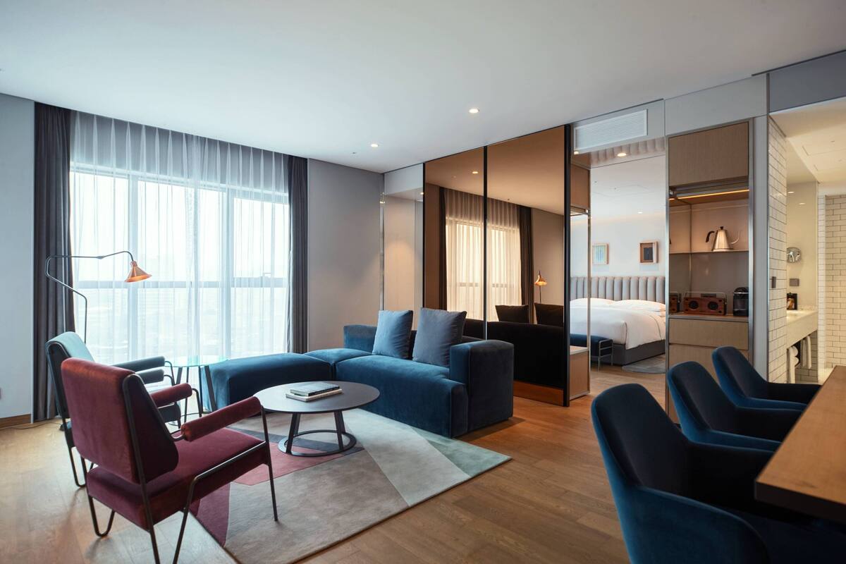 A modern hotel room with a sofa, armchairs, a dining table, and a kitchen area.