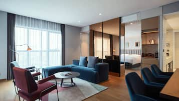 A modern hotel room with a sofa, armchairs, a dining table, and a kitchen area.