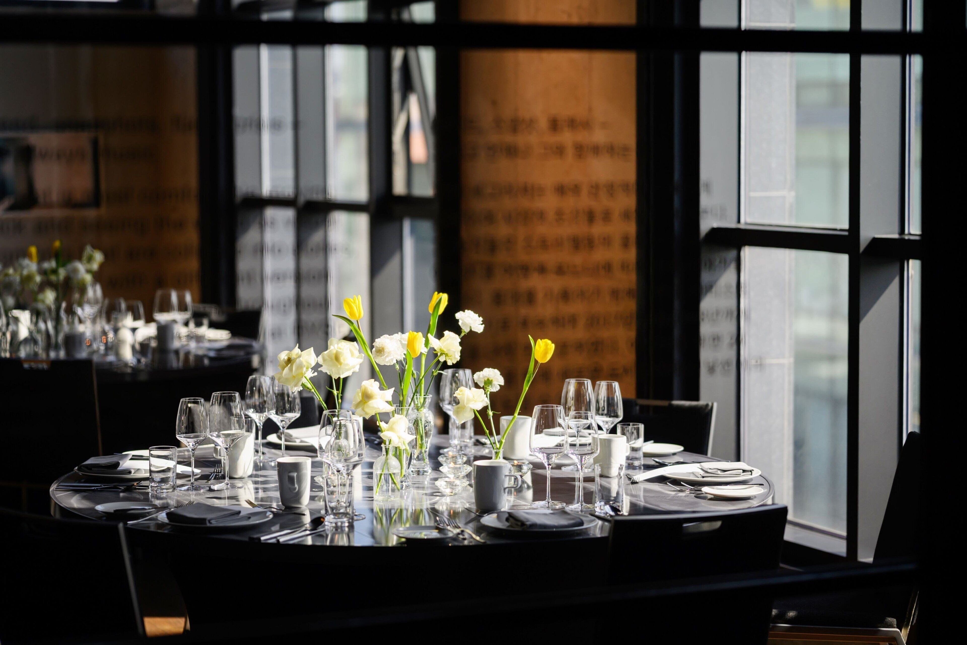 A dining table set with white flowers, clear glasses, and plates, with a view of a building through large windows.