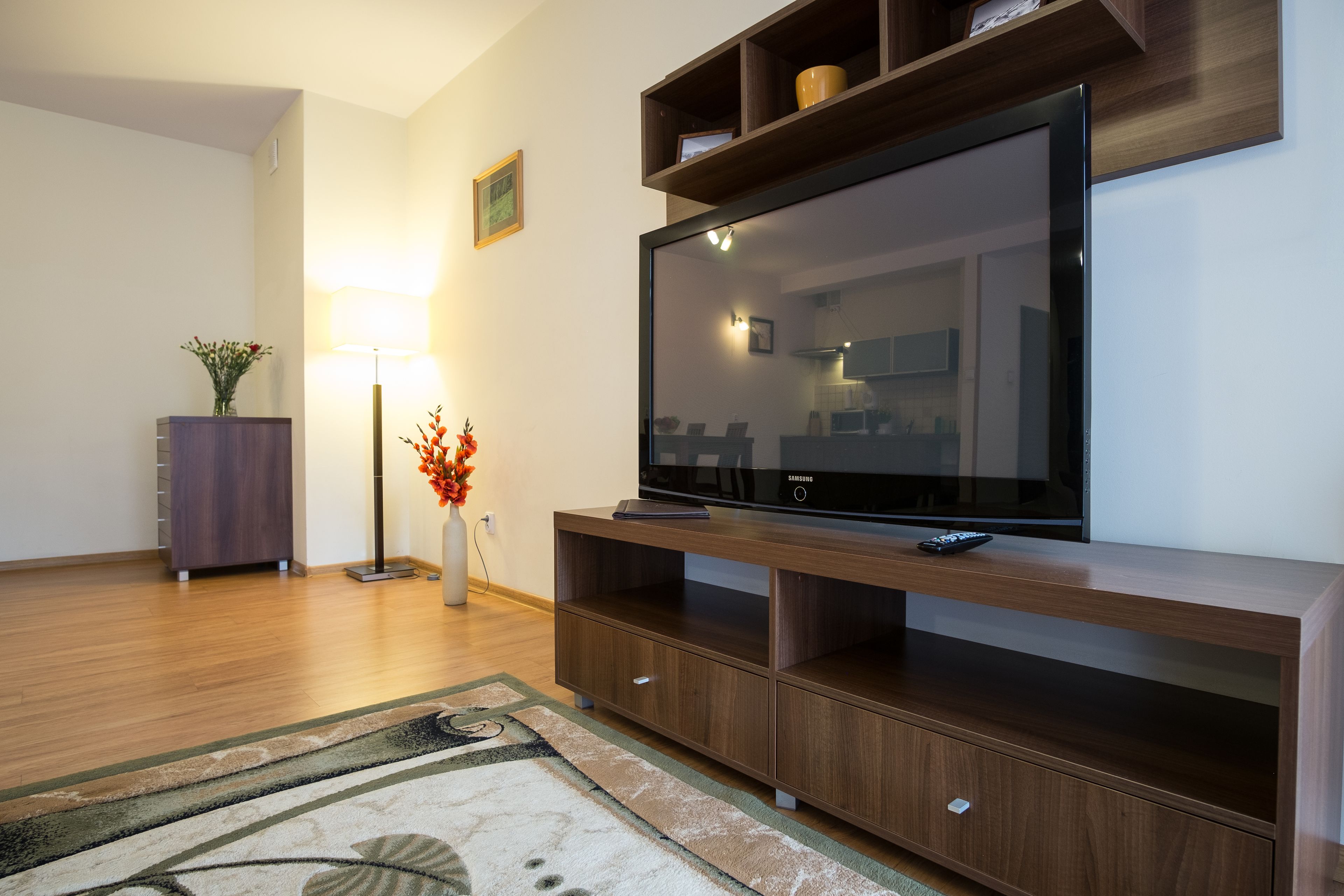A modern living room with a flat-screen TV on a wooden stand, a floor lamp, and a vase with flowers.