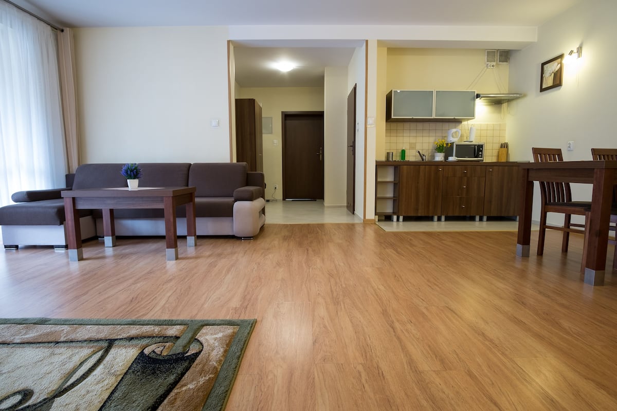 A living room with a sofa, coffee table, and rug. A kitchen is visible in the background with cabinets and a microwave.