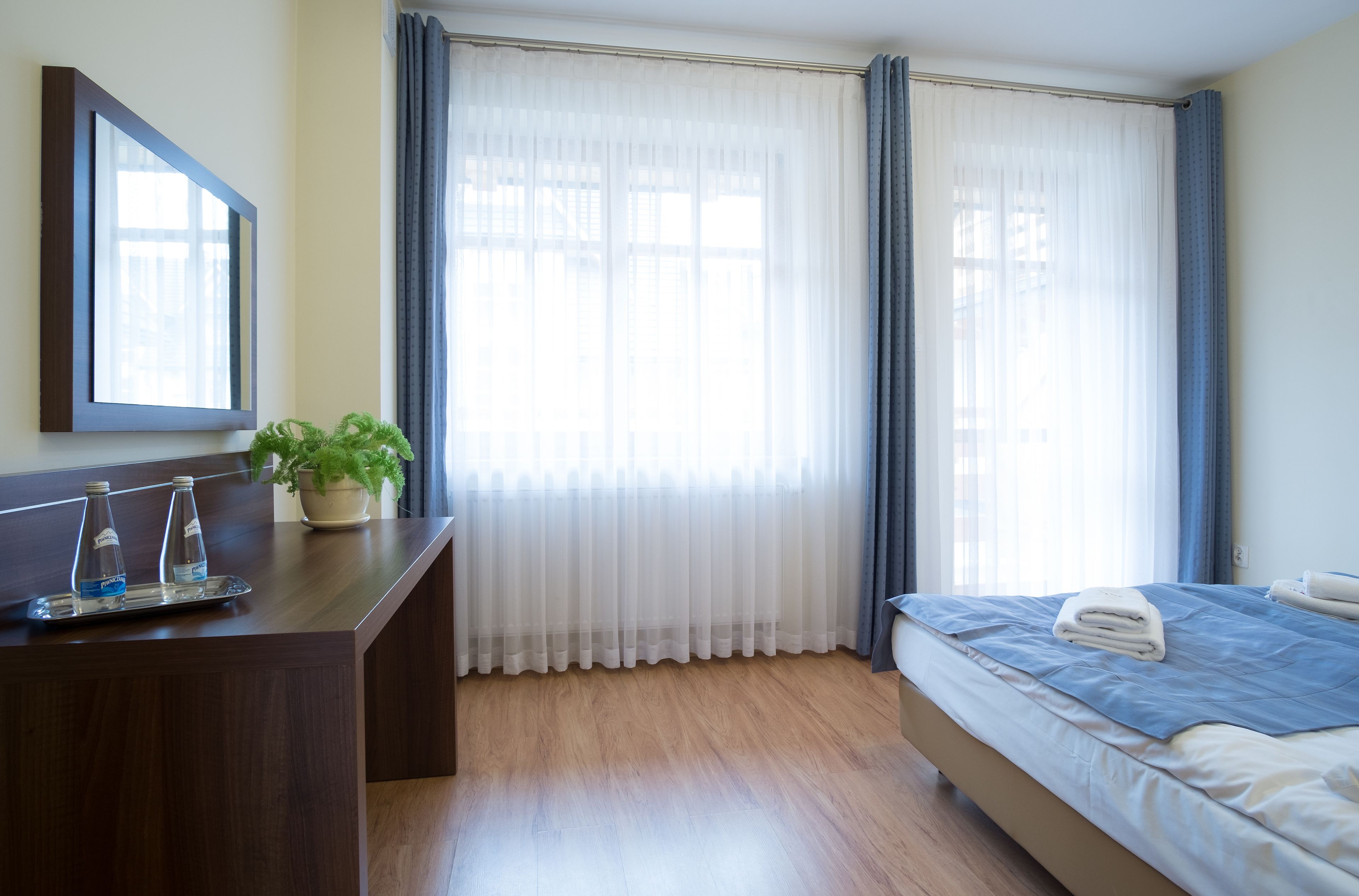 Habitación de hotel con escritorio de madera, planta en maceta, cama con colcha azul y ventana con cortinas traslúcidas.