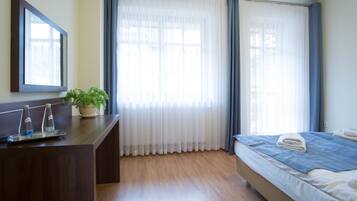 Habitación de hotel con escritorio de madera, planta en maceta, cama con colcha azul y ventana con cortinas traslúcidas.