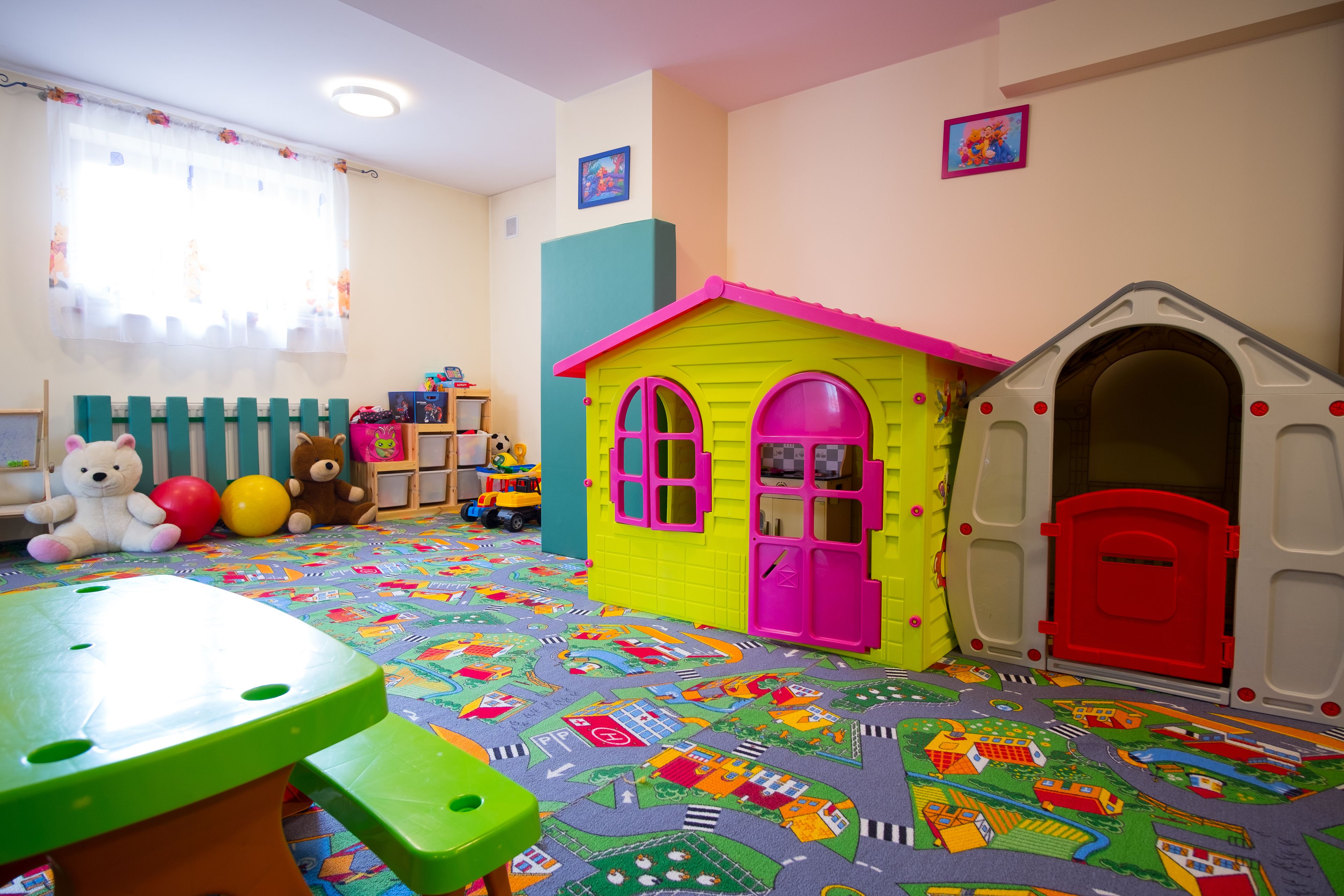 Uma sala de jogos com uma casinha colorida, uma mesa verde e um tapete de atividades com diversos desenhos.