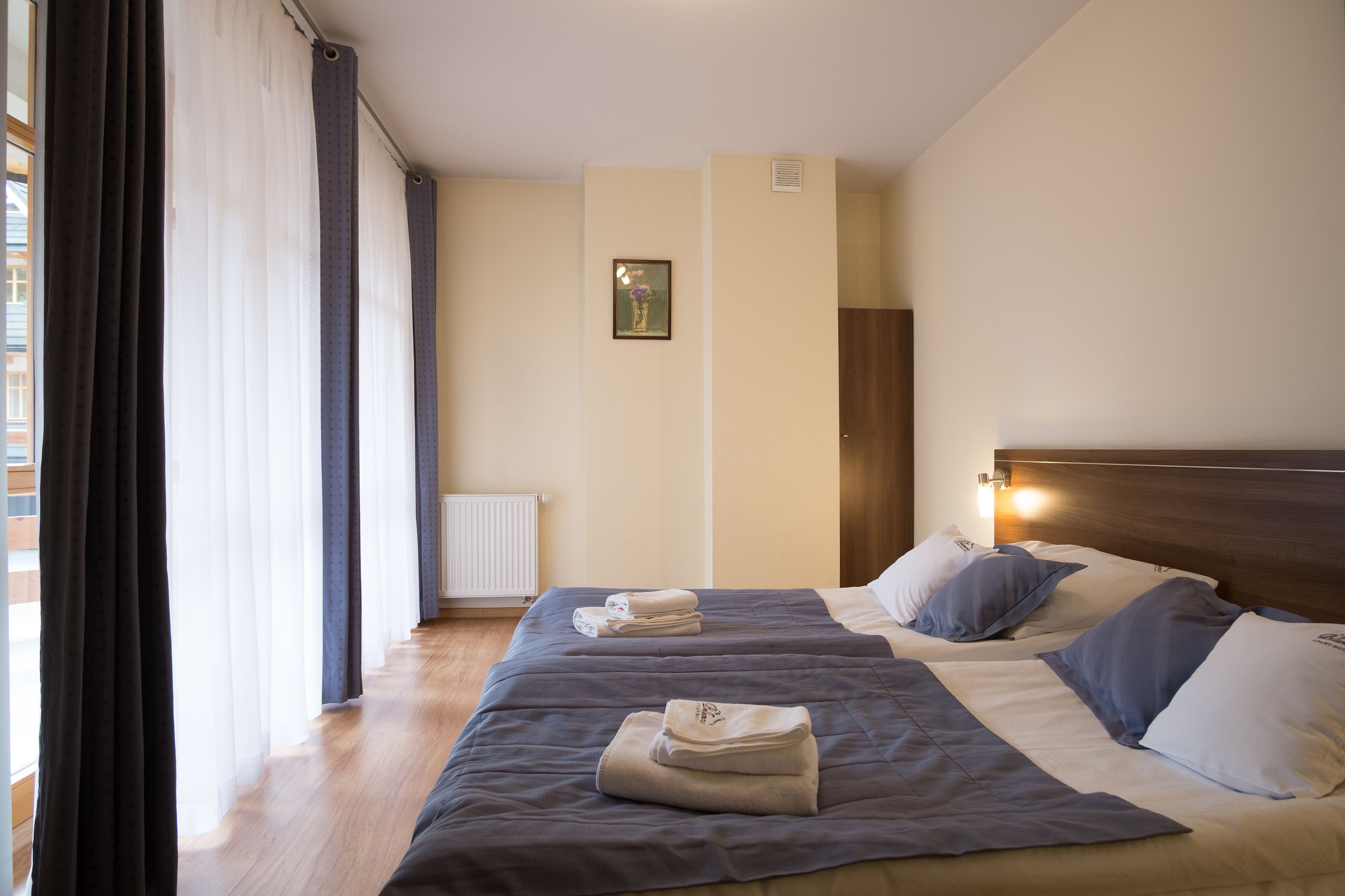 A hotel room with two beds, a radiator, a framed picture on the wall, and a window with curtains.