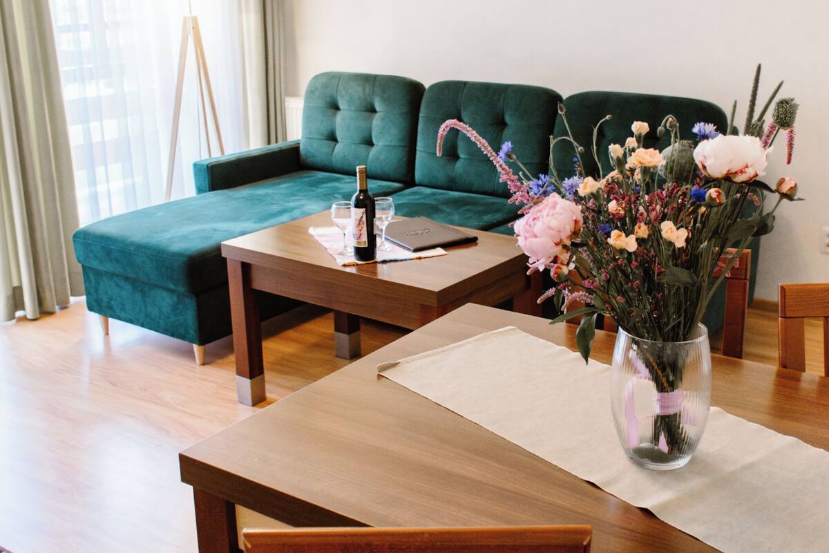 A modern living room with a green sofa, wooden dining table, and a vase of flowers.