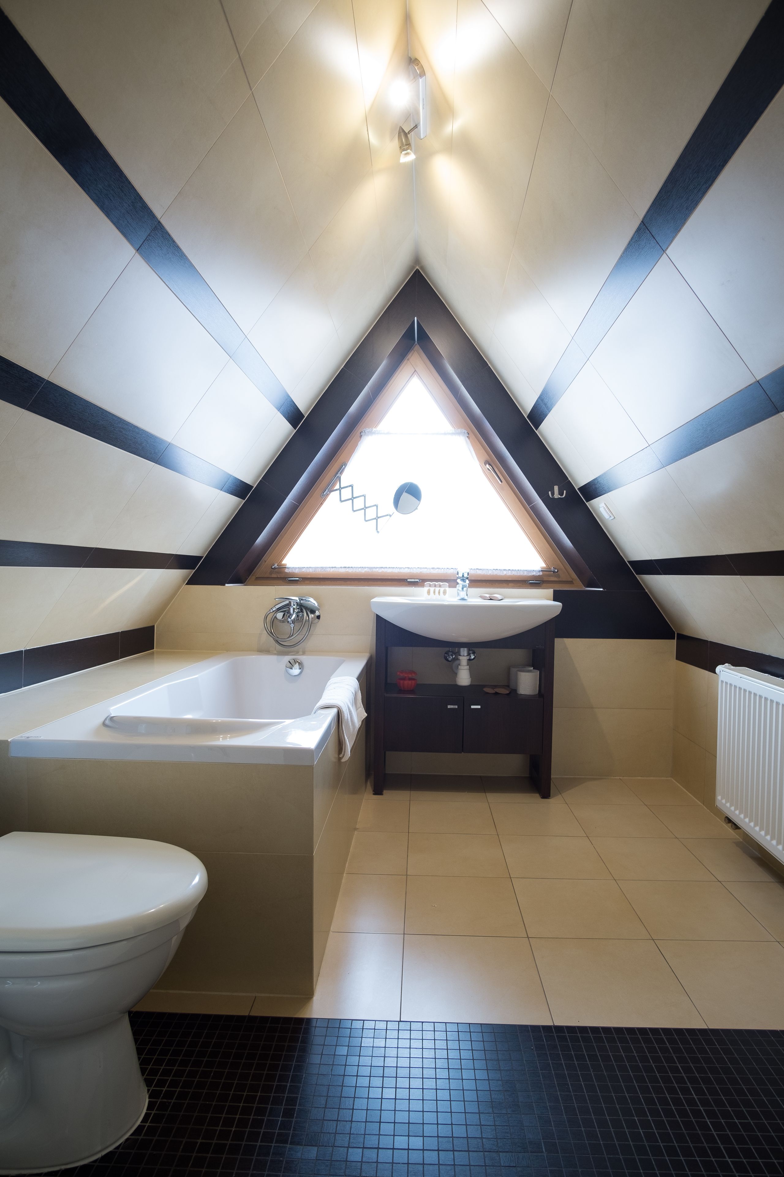 Un baño con bañera, inodoro y lavamanos bajo un techo inclinado con tragaluz.
