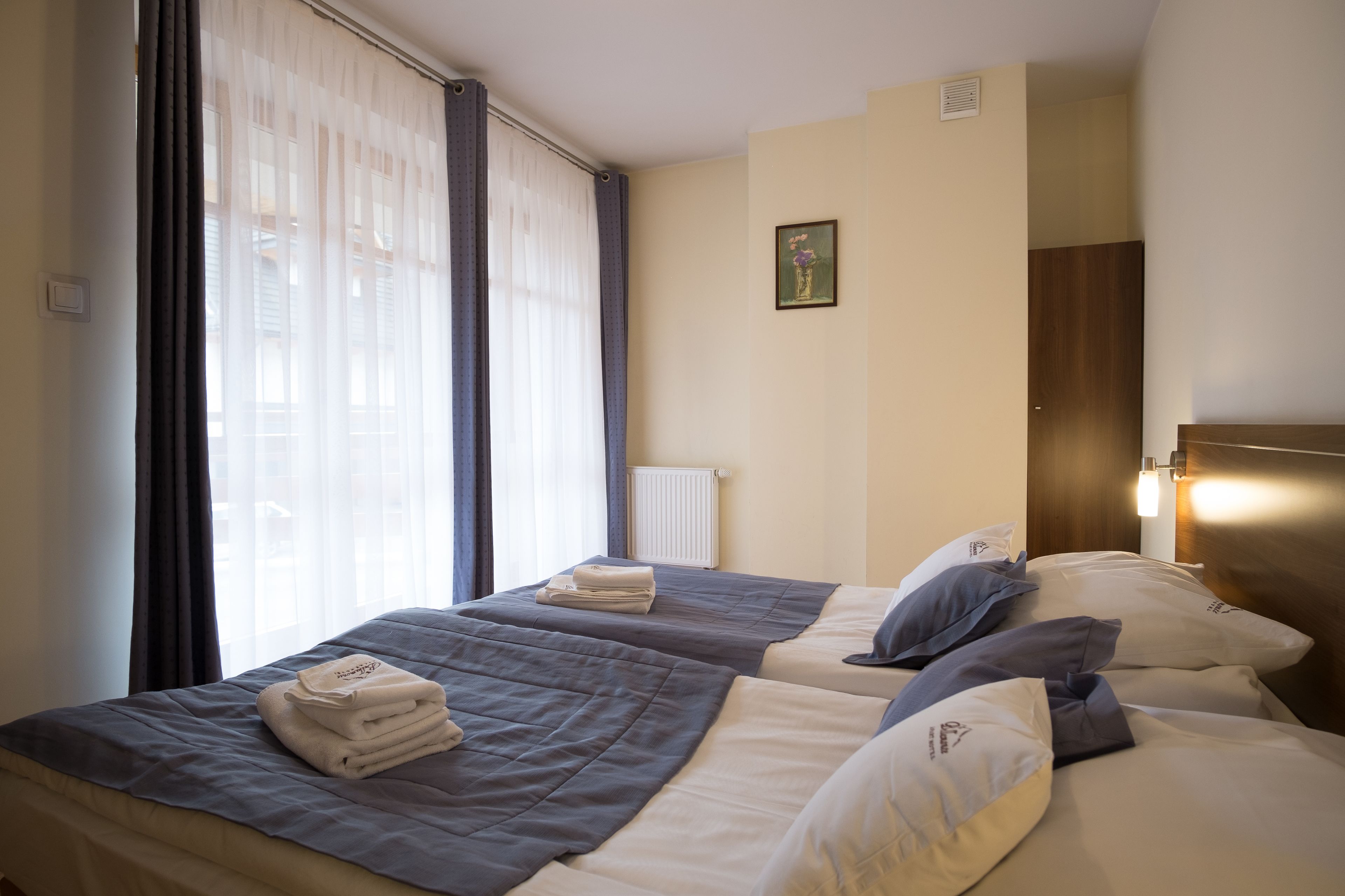 A hotel room with two beds, a window with curtains, a radiator, and a framed picture on the wall.
