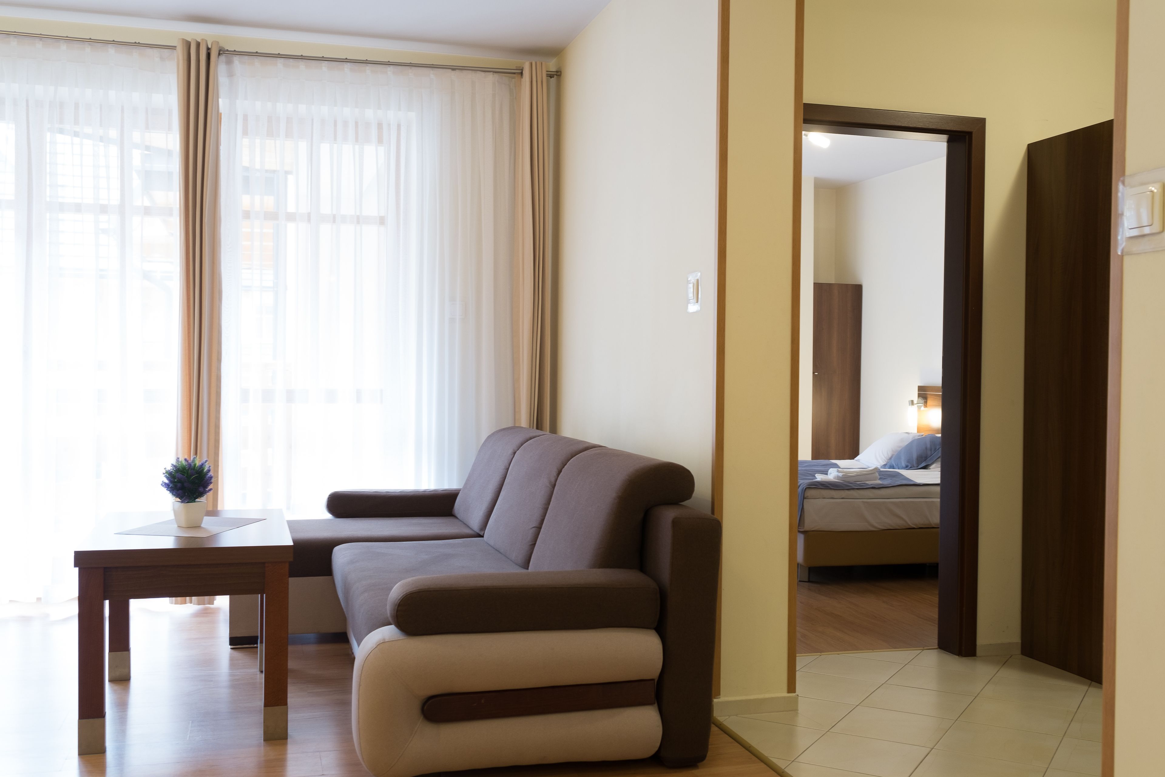 A modern living room with a sofa, a small table, and a potted plant. A bedroom is visible through an open door.