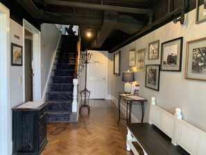 A hallway with a wooden floor, a staircase with patterned carpet, a lamp, and framed pictures on the walls.