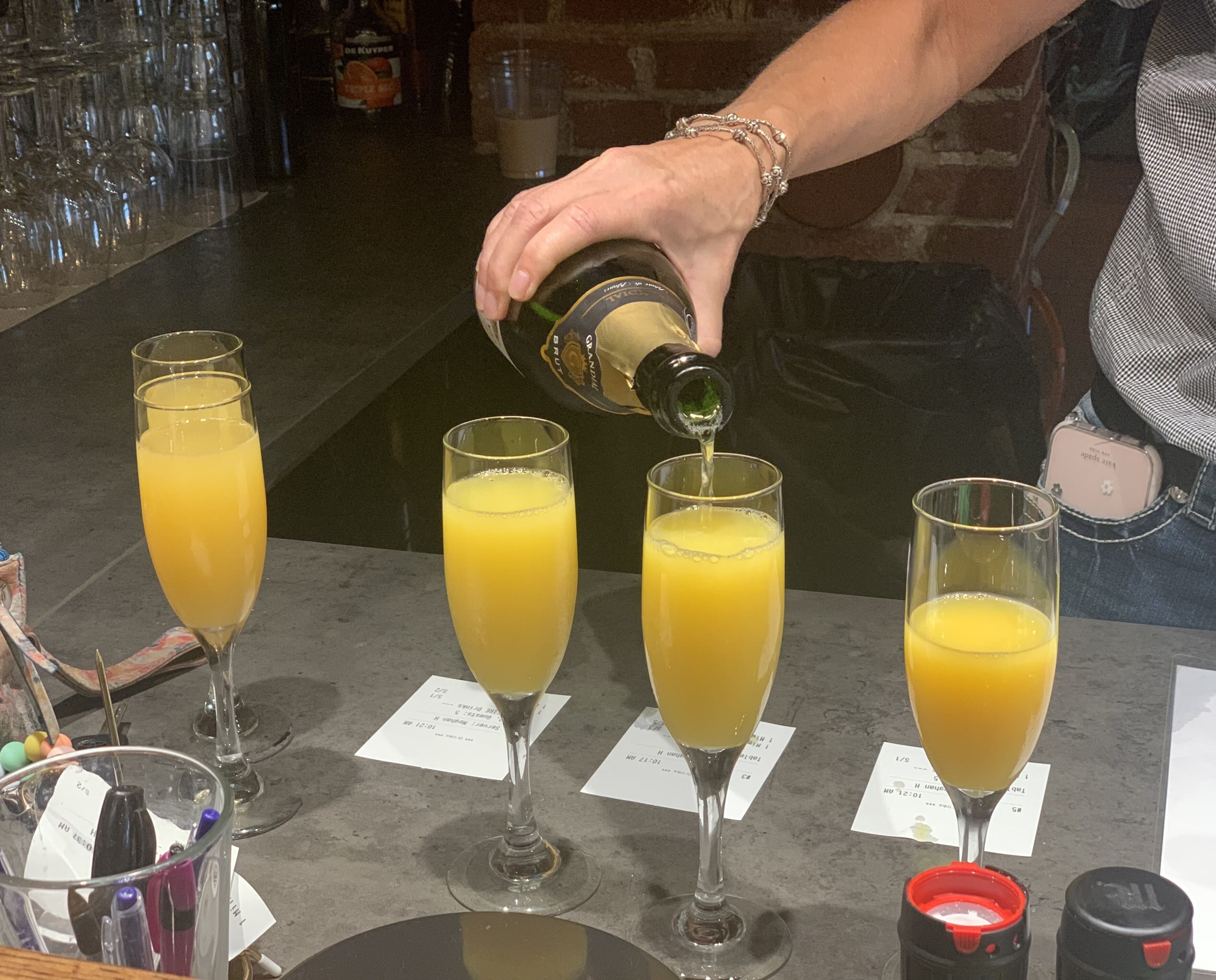 A bartender pouring orange juice into four champagne flutes on a bar counter.