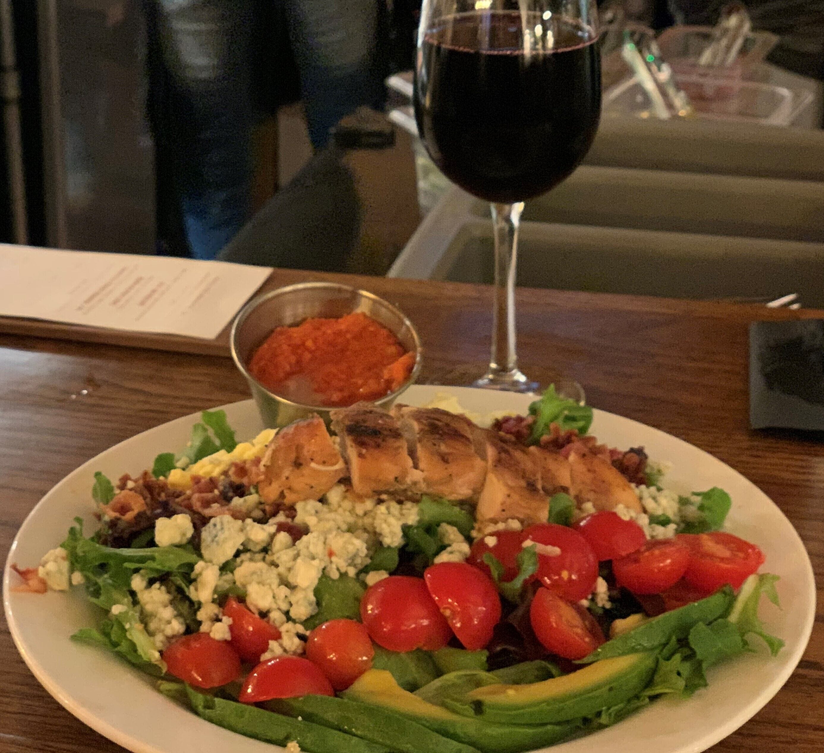 A plate of food with grilled chicken, avocado, and cherry tomatoes on a bed of greens, accompanied by a glass of red wine and a small bowl of sauce.