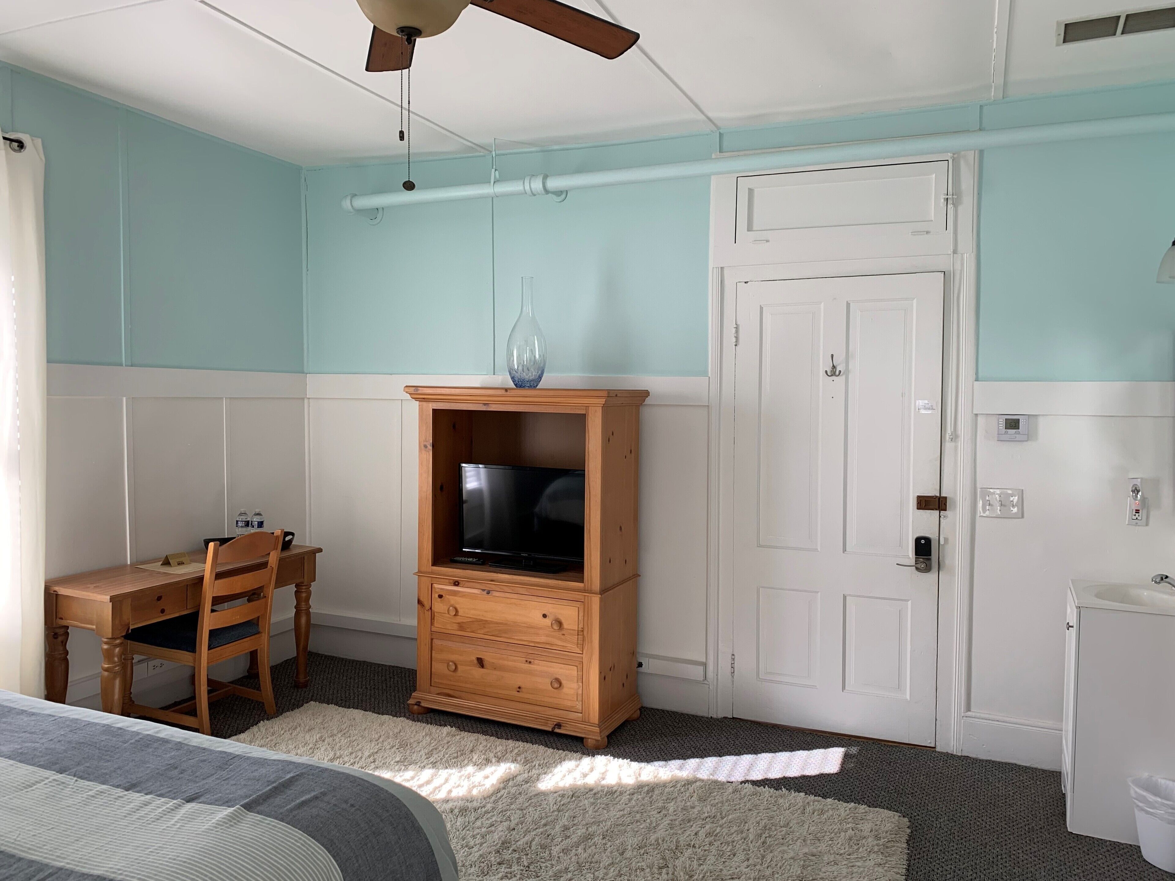 A bedroom with a bed, a wooden TV stand, a desk with a chair, a white door, and a small white cabinet.