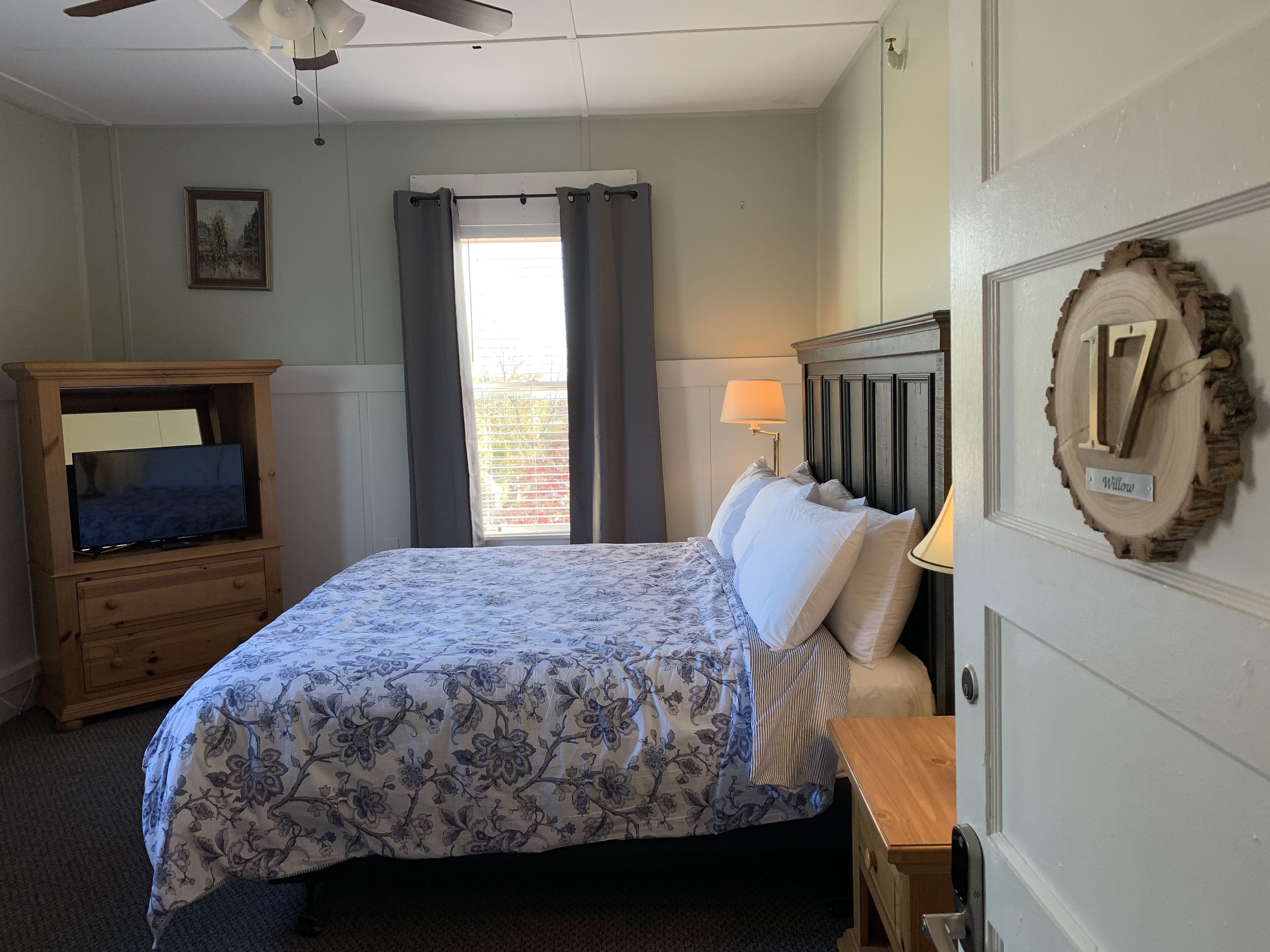 A bedroom with a bed, a wooden dresser, a ceiling fan, a window with curtains, a lamp, and a wall-mounted number plate.