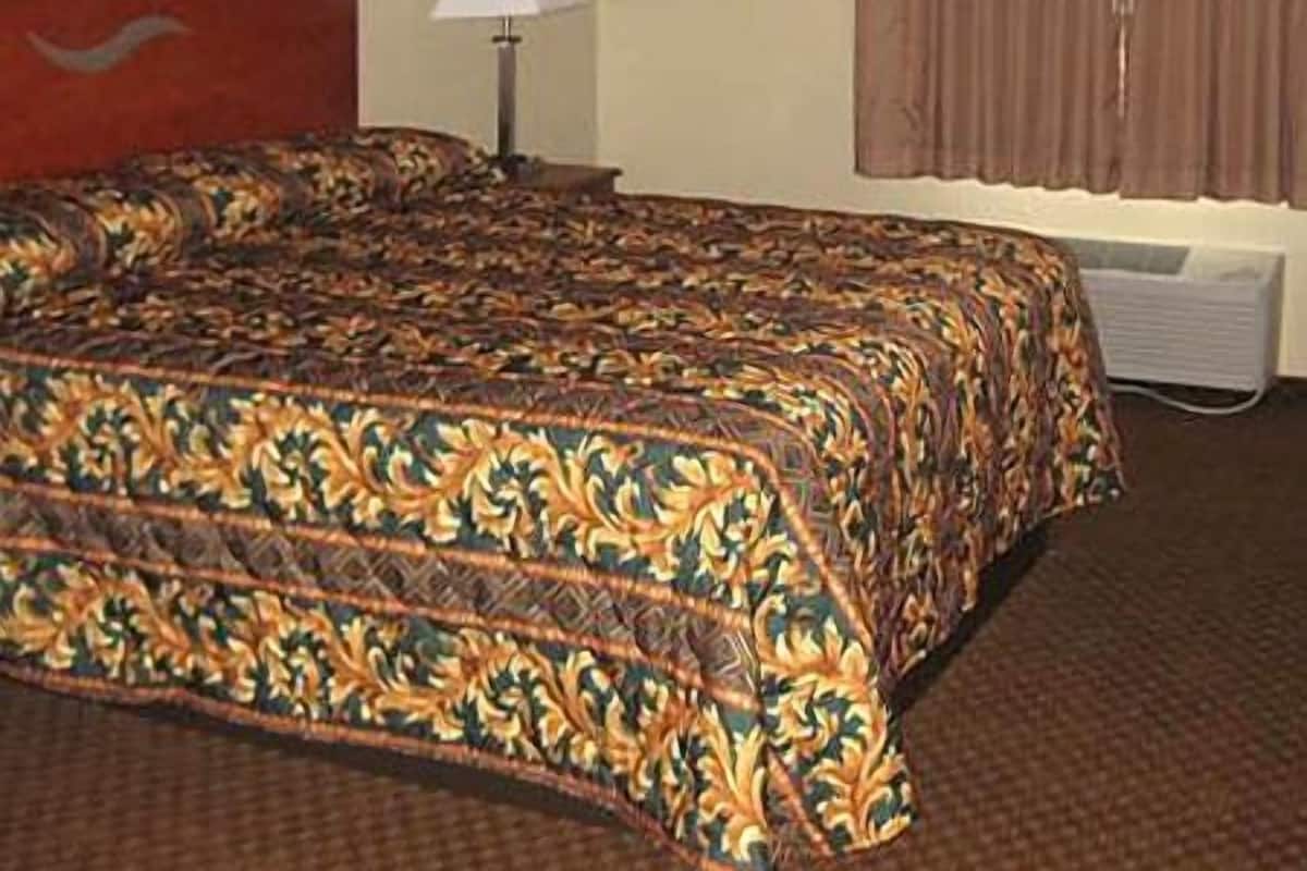Quarto de hotel com uma cama grande, uma luminária, uma janela com cortinas e uma pintura na parede.