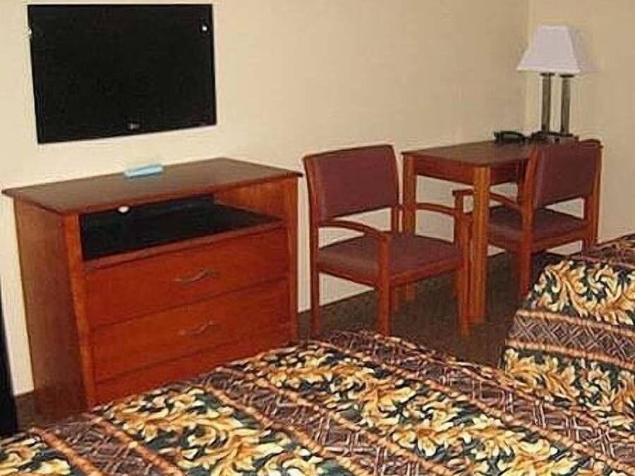A hotel room with a bed, a desk with drawers, a television, two chairs, and a table with a lamp.