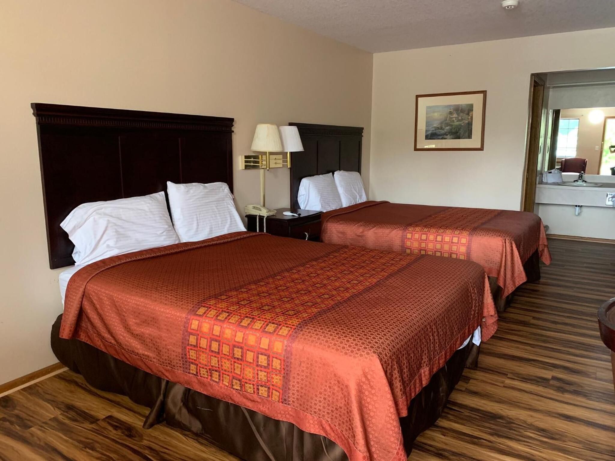 A hotel room with two beds, a nightstand, a lamp, a framed picture on the wall, and a bathroom visible through an open door.