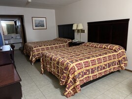 Habitación de hotel con dos camas, un escritorio, un tocador, una mesita de noche, una lámpara y un cuadro en la pared.