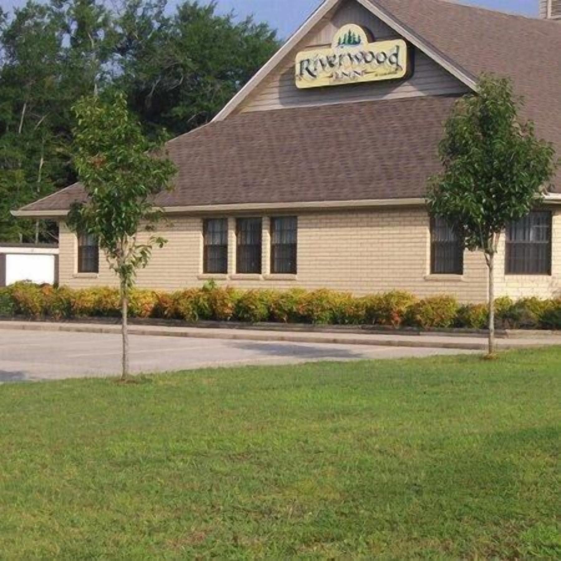 A two-story building with a sign that reads "Riverwood Inn." There are three windows on the front facade, and the building is surrounded by a well-maintained lawn and a row of bushes.