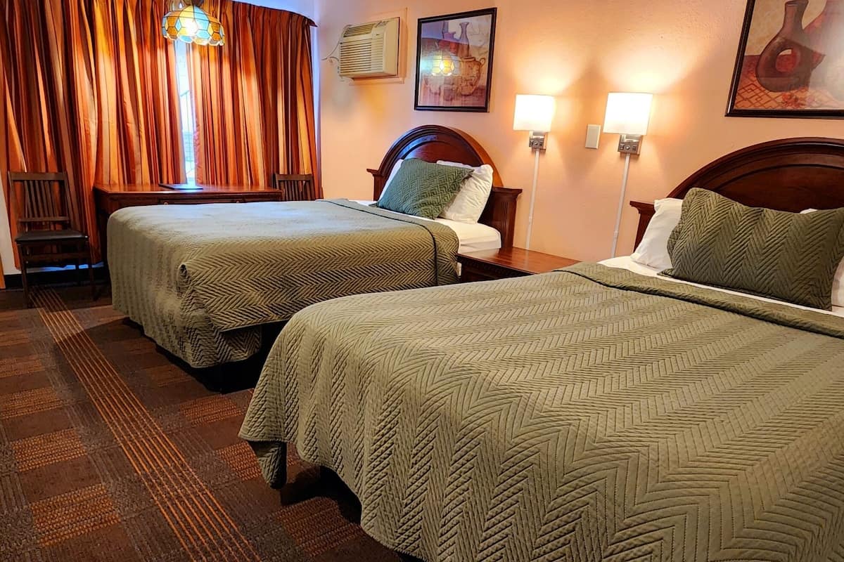 Une chambre d’hôtel avec deux lits, une tête de lit en bois, un bureau, une chaise, une fenêtre avec des rideaux, des lampes fixées au mur et des œuvres d’art.