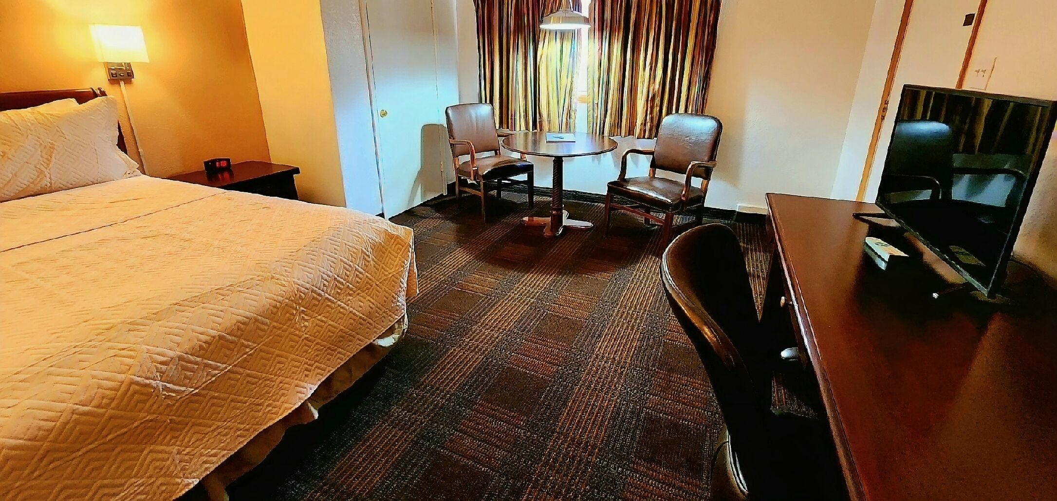 A hotel room with a bed, a small table with chairs, a TV, and a window with curtains.