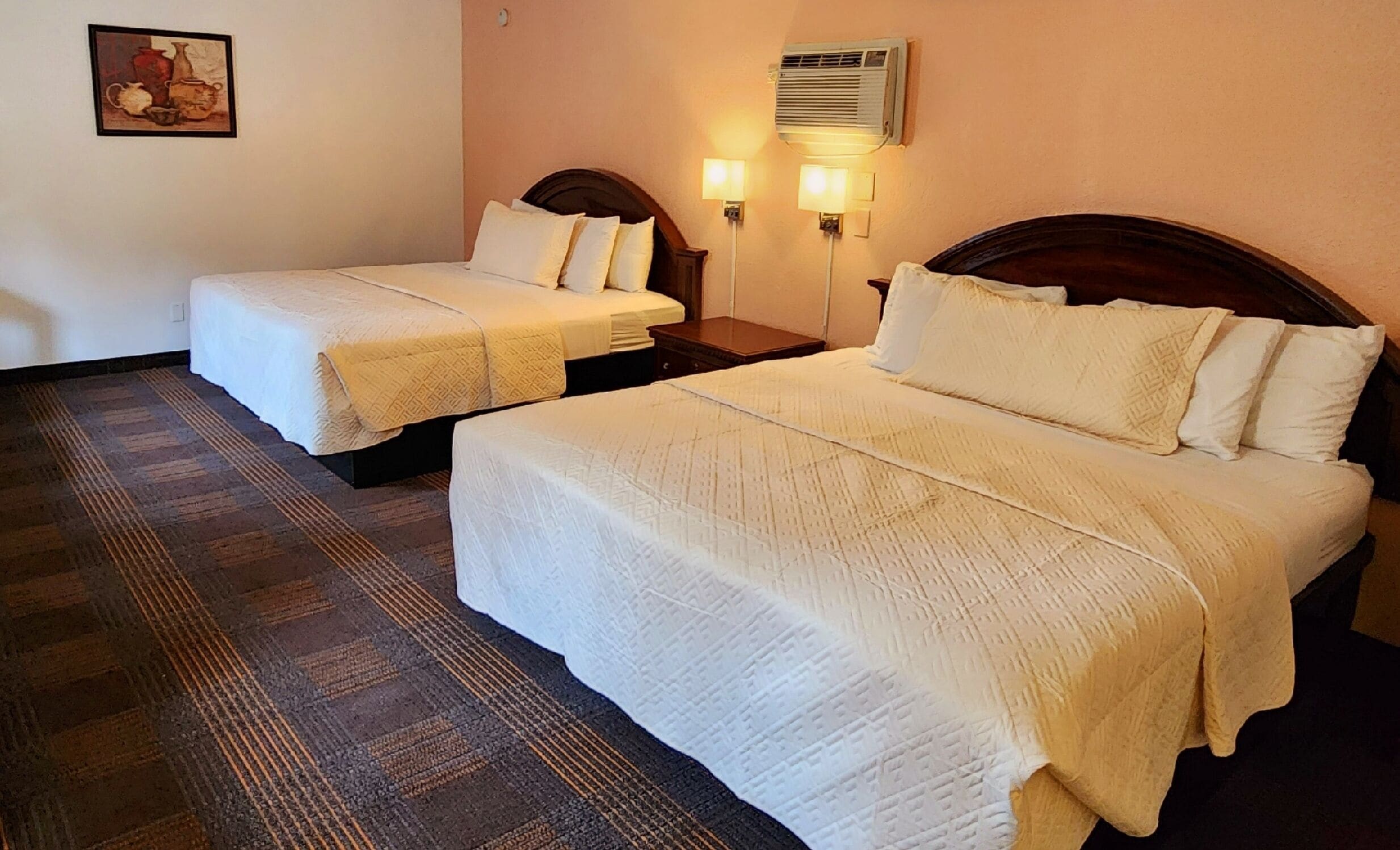 Dos camas individuales con ropa de cama blanca, una mesita de noche pequeña, un aire acondicionado de pared y una obra de arte enmarcada en la pared.
