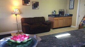 A living room with a brown sofa, a glass coffee table with a flower, a lamp, and a kitchen area with wooden cabinets and a countertop.