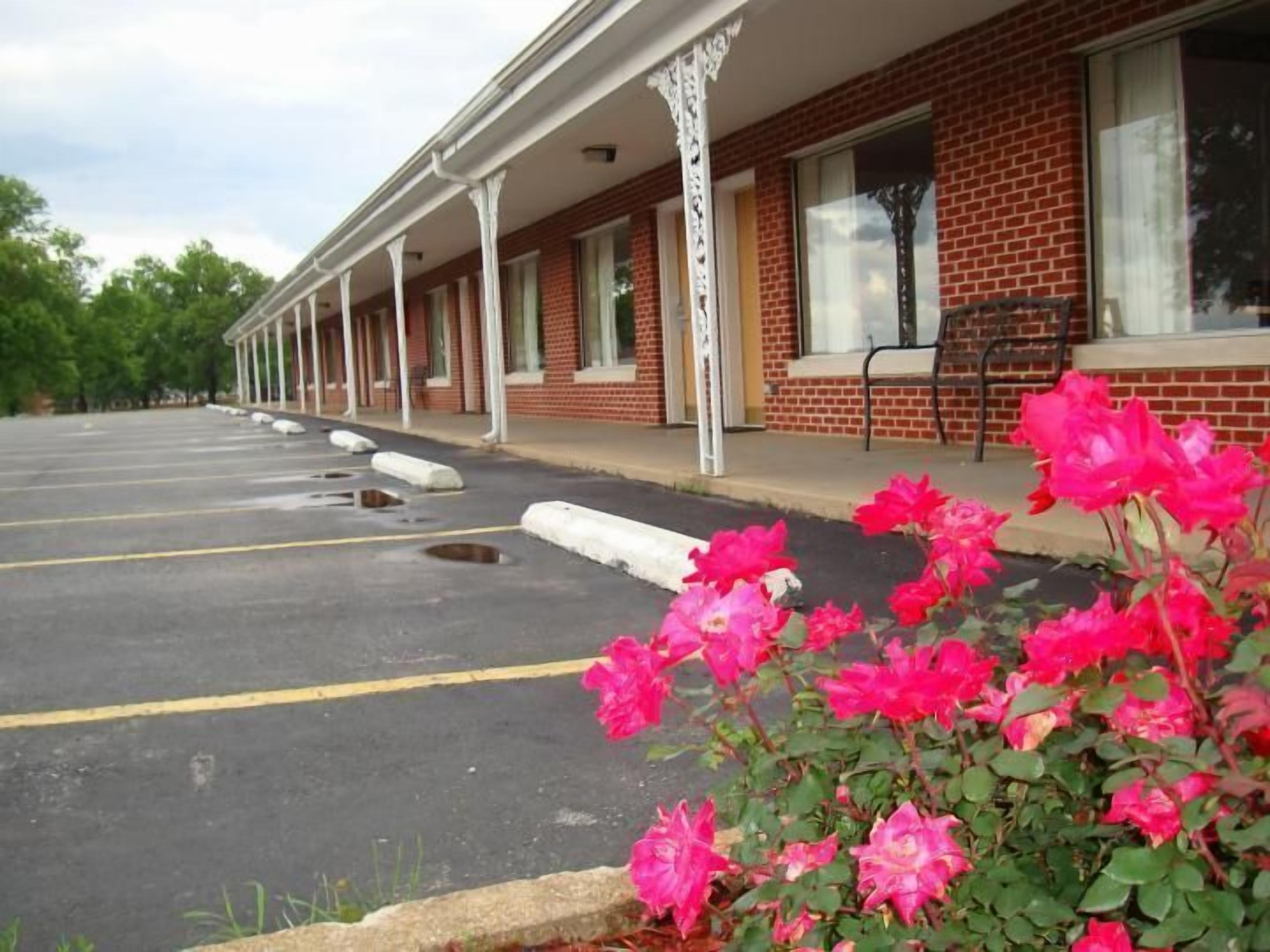 A motel with a row of rooms, a parking lot, and a row of pink flowers in the foreground.