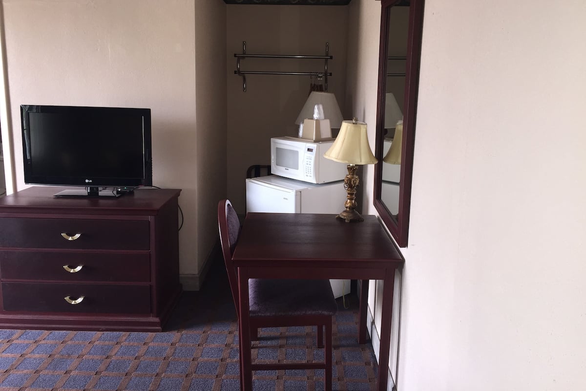 A hotel room with a TV on a wooden dresser, a microwave, a lamp, and a chair.