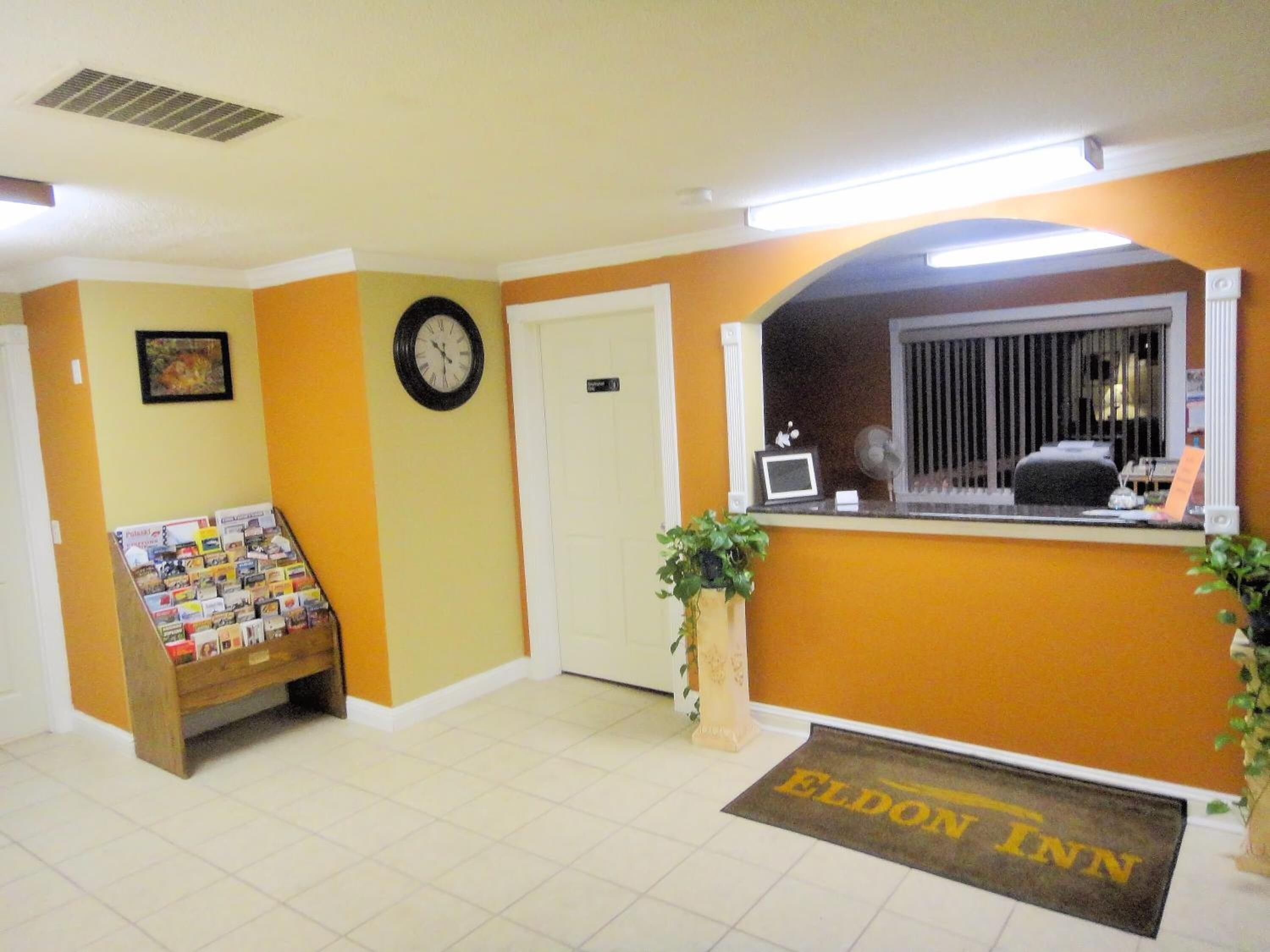 A hotel reception area with a clock, a desk with a computer, a plant, and a welcome mat.