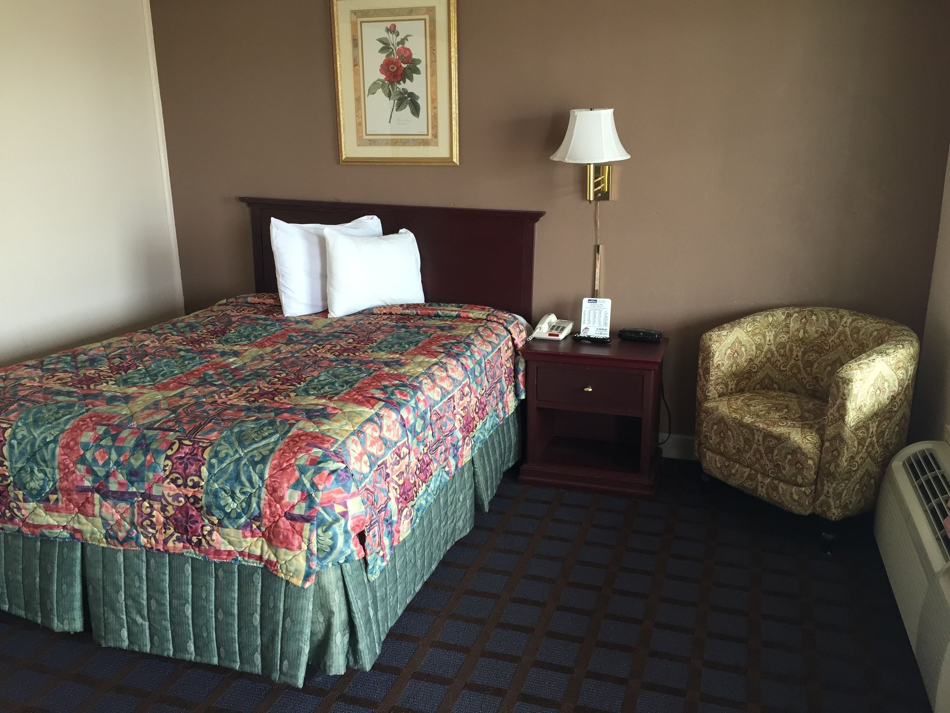 A hotel room with a bed, a chair, a nightstand with a lamp, and a framed picture on the wall.