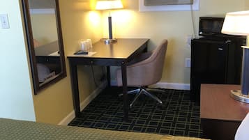 A hotel room with a bed, a desk with a chair, a lamp, a television, and an air conditioning unit.