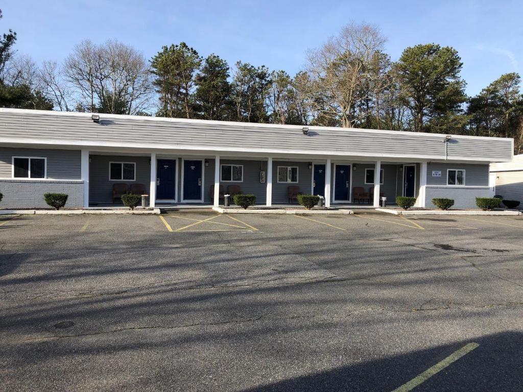 Un motel con una hilera de habitaciones, un estacionamiento y árboles al fondo.