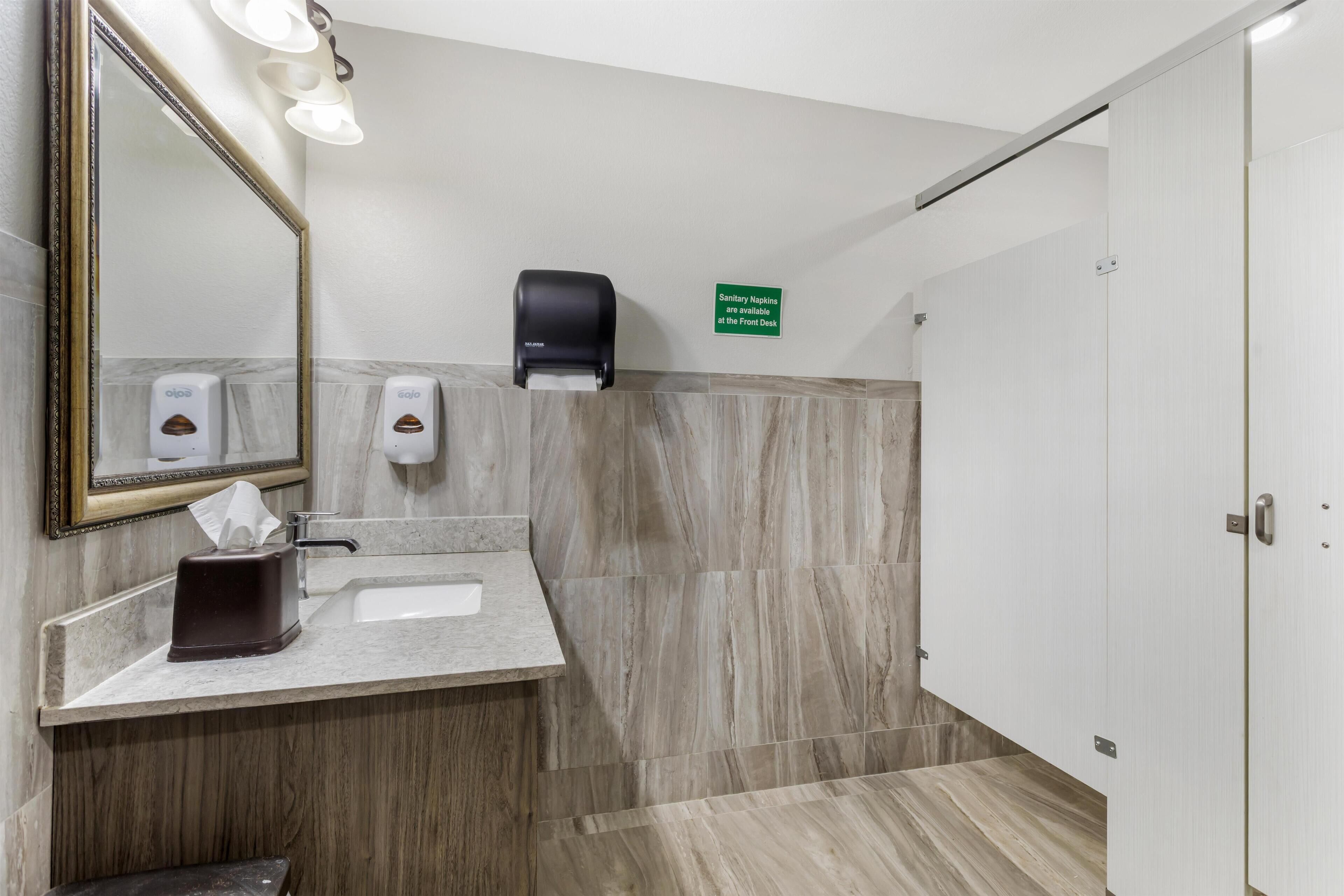 Casa de banho moderna com lavatório de mármore, dispensador de lenços de papel, secador de mãos e aviso a indicar a disponibilidade de pensos na receção.