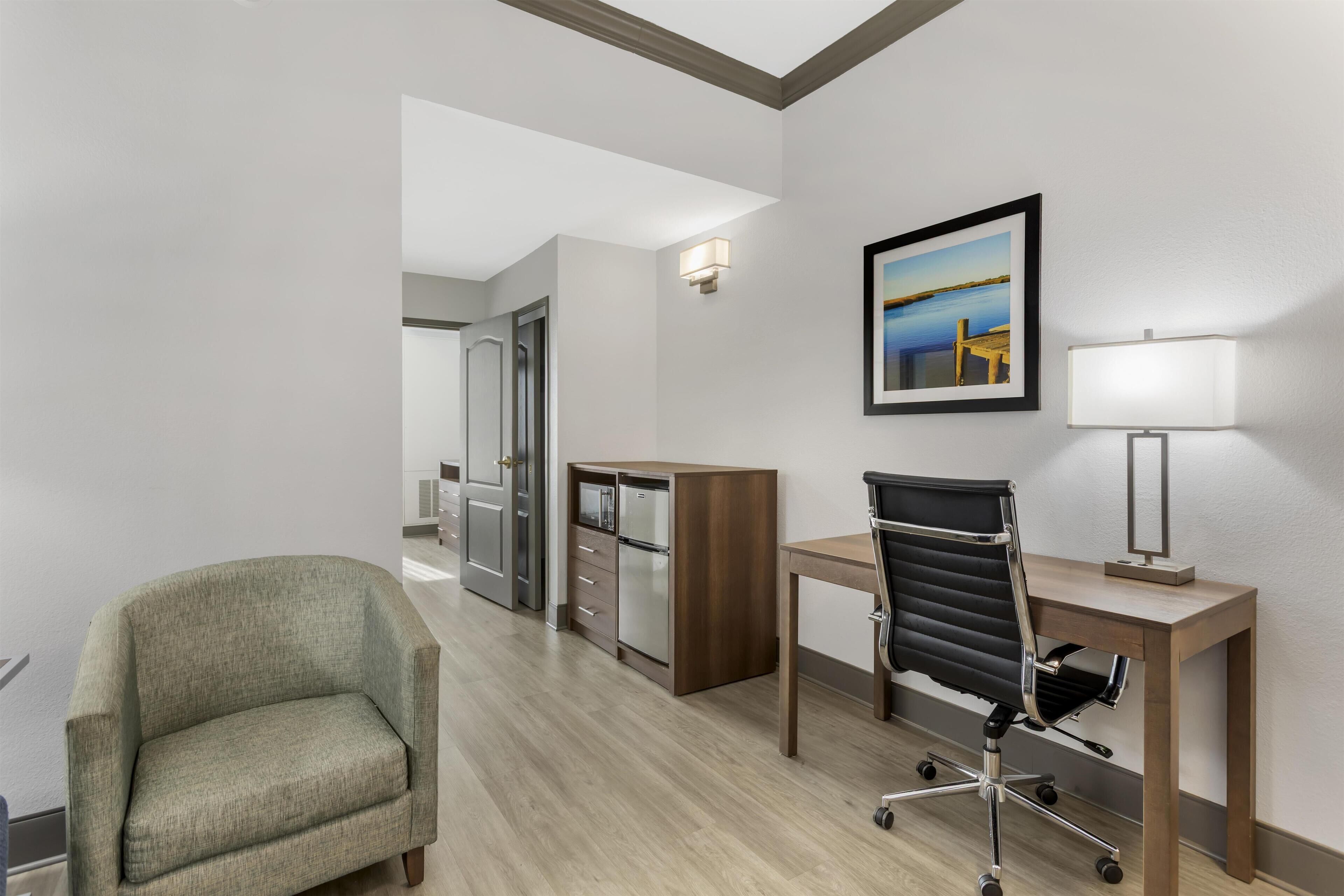 A hotel room with a green armchair, a desk with a black office chair, a wooden cabinet, a framed picture on the wall, and a lamp.