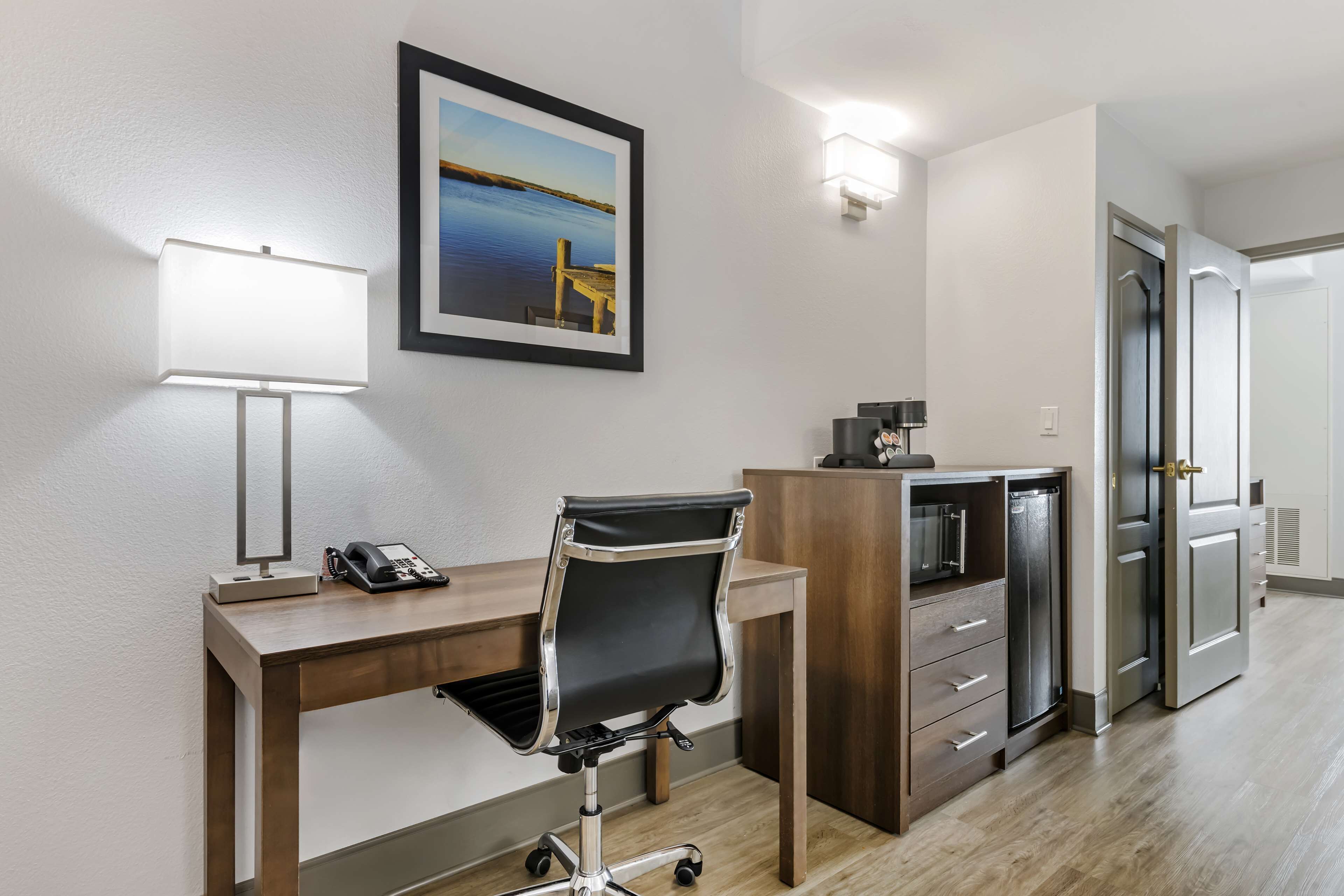 Habitación de hotel con escritorio, silla y refrigerador. Se observa un cuadro en la pared y una puerta a la derecha.