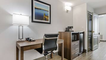 Habitación de hotel con escritorio, silla y refrigerador. Se observa un cuadro en la pared y una puerta a la derecha.