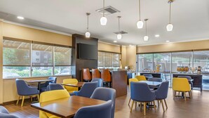 Un comedor moderno con mesas y sillas de madera, ventanas amplias y una encimera de cocina visible a través del vidrio.