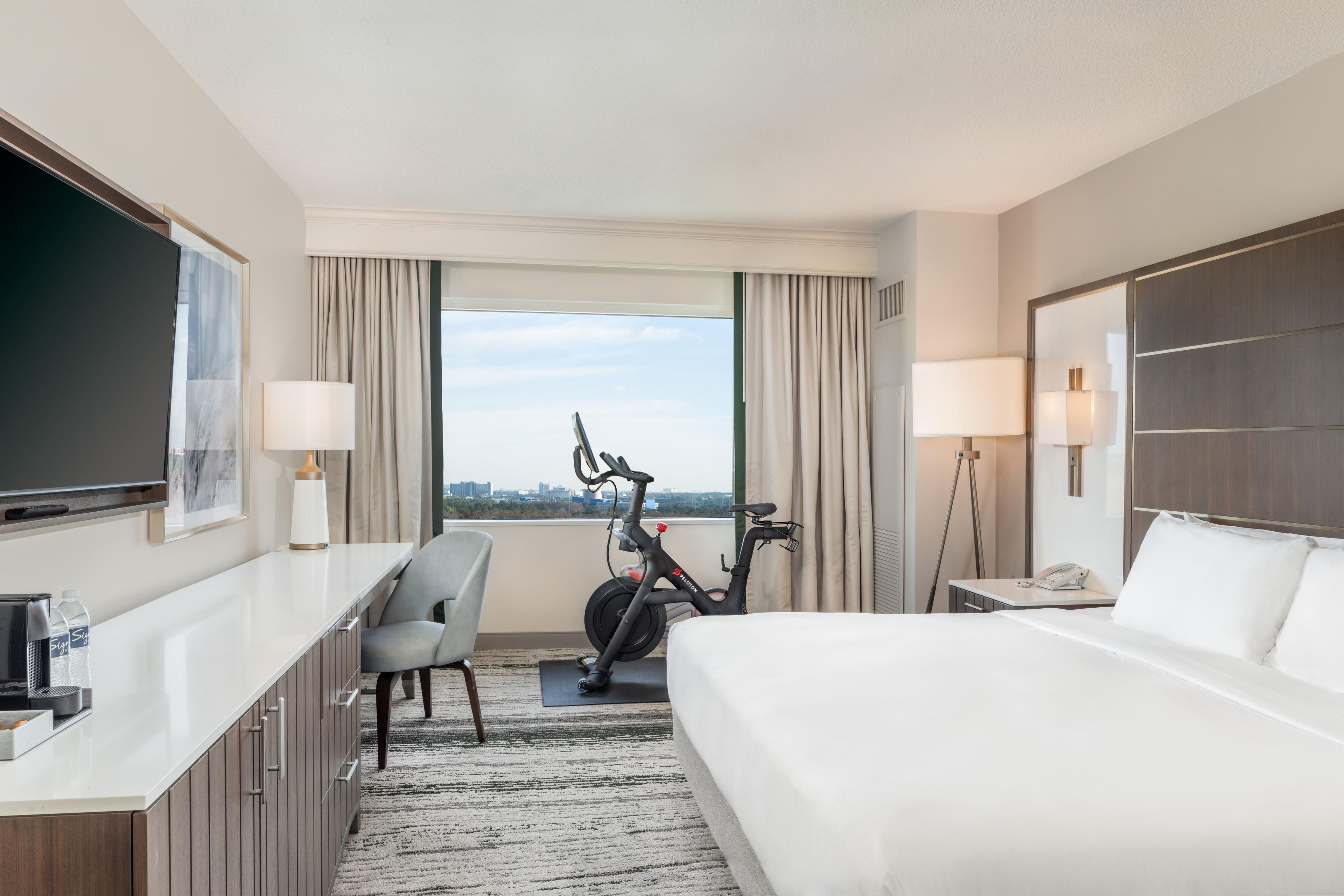 A hotel room with a bed, a desk, a chair, a TV, a bike, and a view of the city.