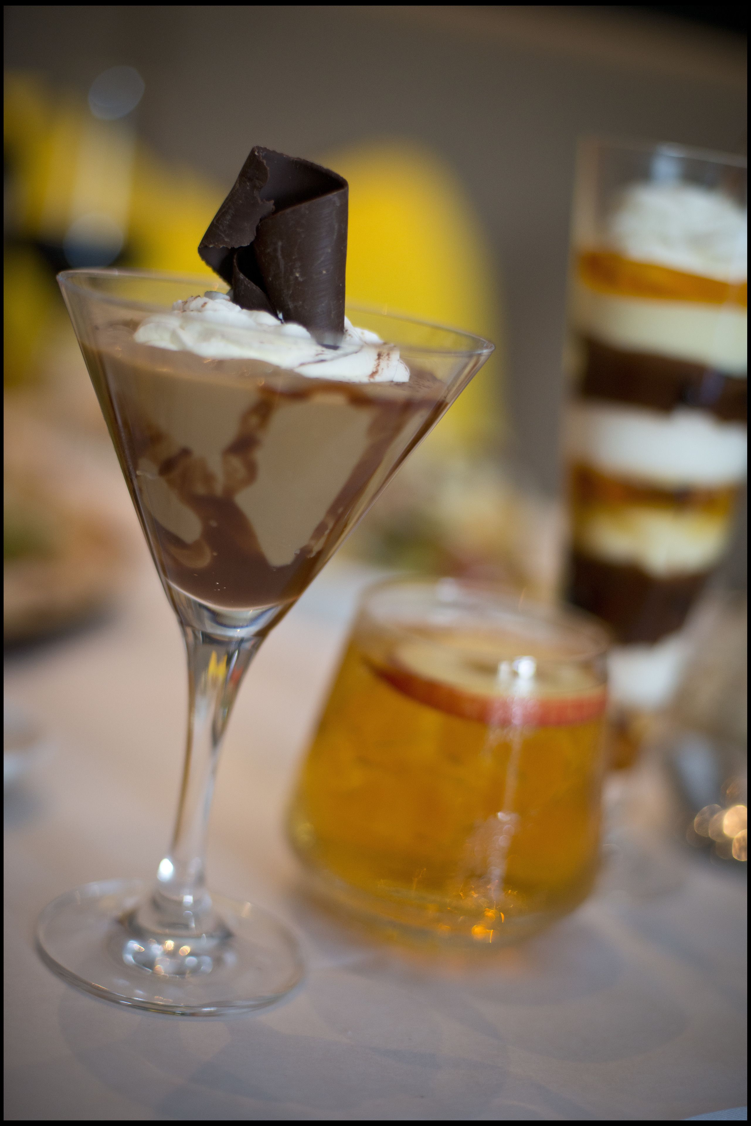 A dessert in a martini glass with whipped cream and chocolate, and a glass of apple cider in the background.