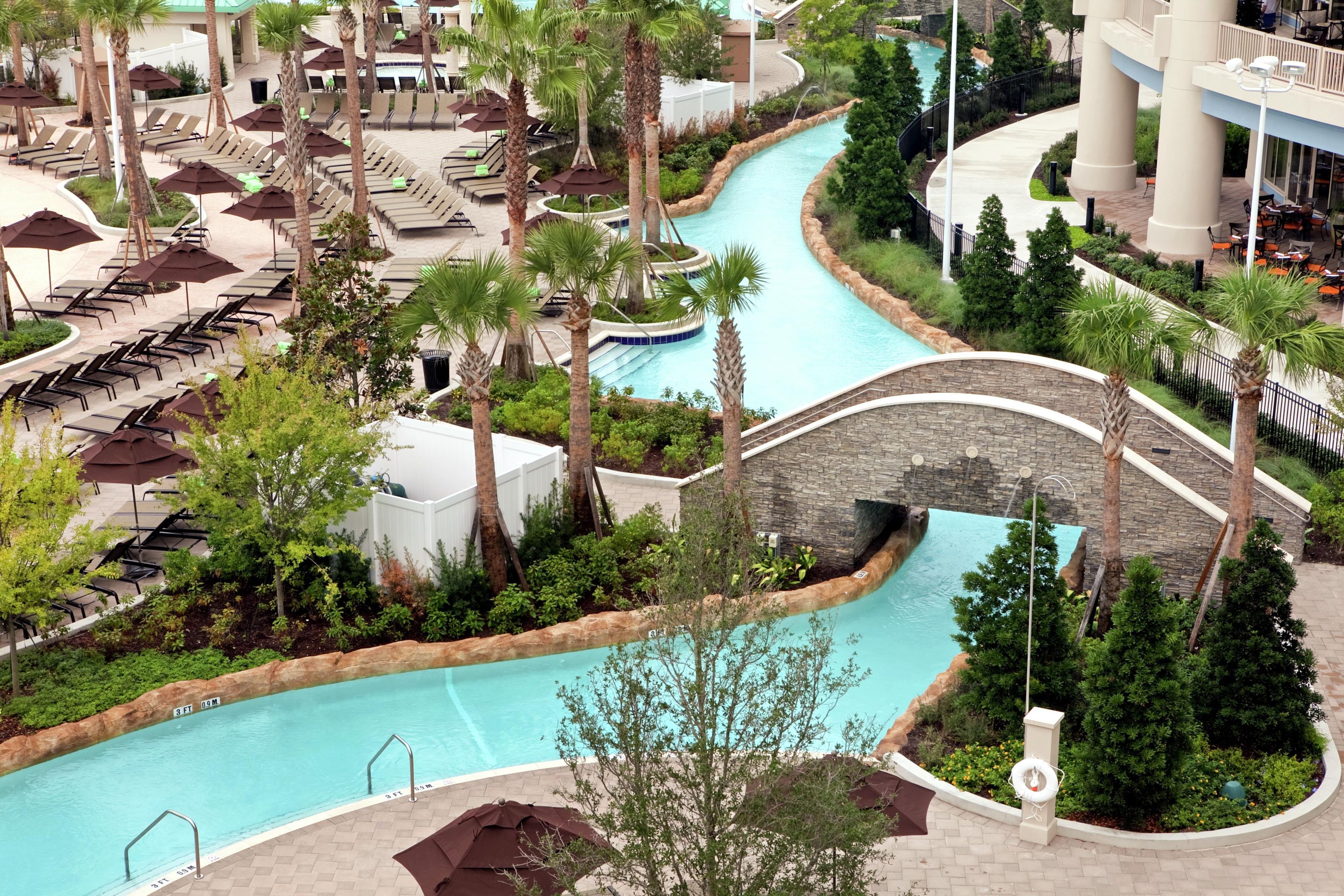 A resort with a pool, lounge chairs, palm trees, and a stone bridge.