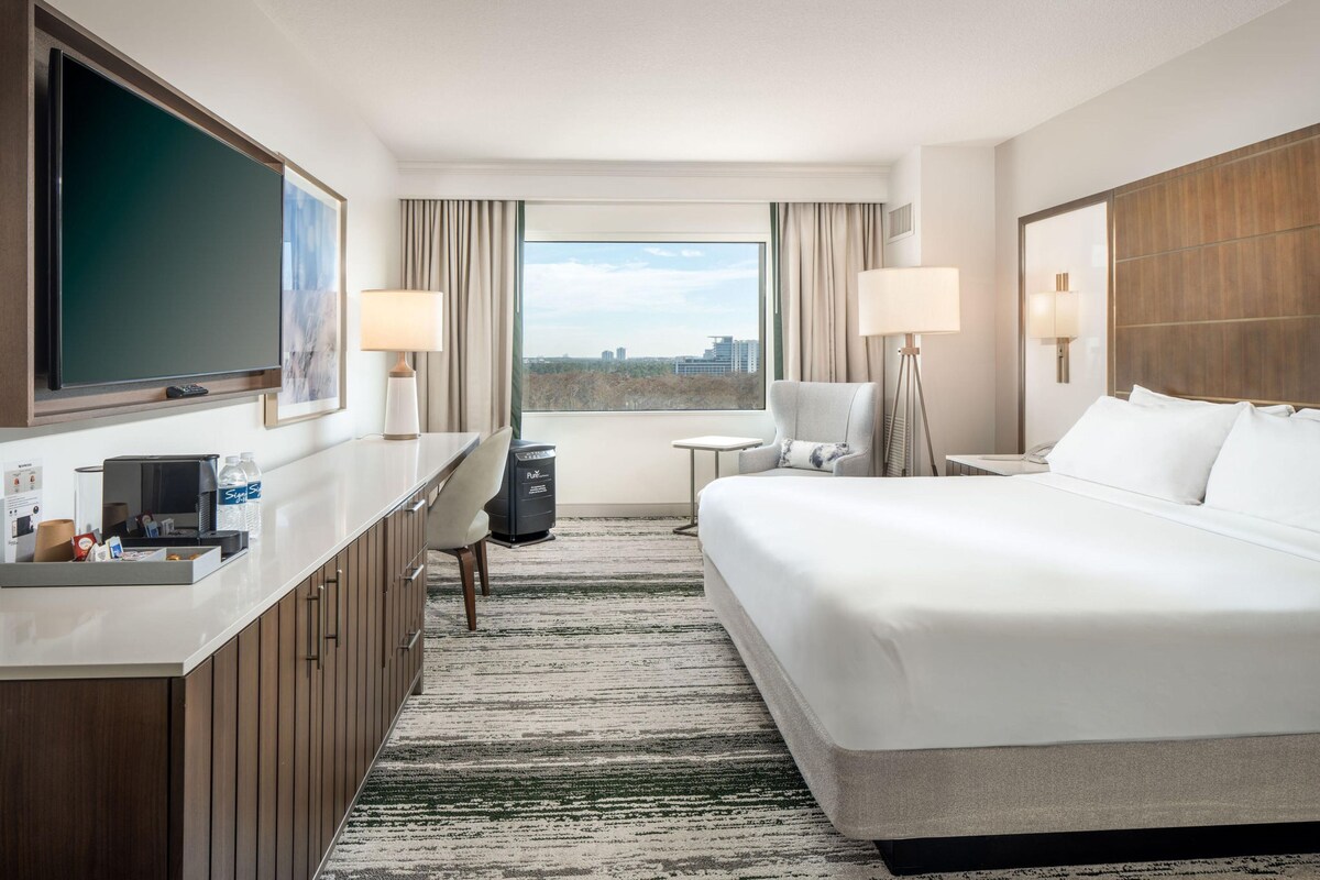 A hotel room with a bed, desk, chair, TV, and a view of the city.