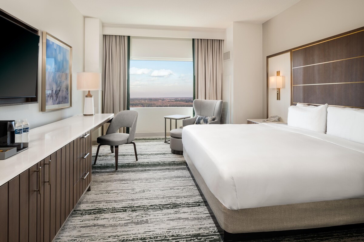 A hotel room with a large bed, a desk with a chair, a TV, and a window with a view.