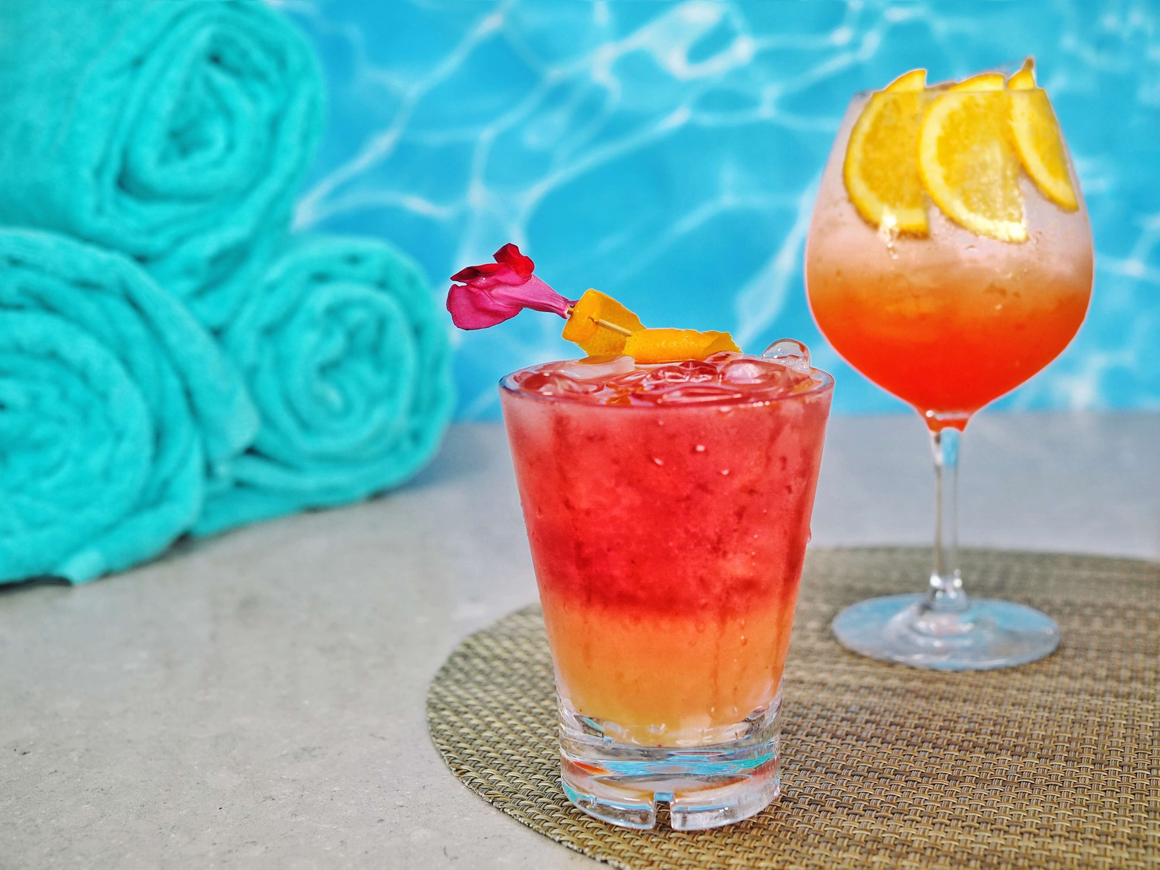 Two cocktails with garnishes, a blue background, and a woven mat.