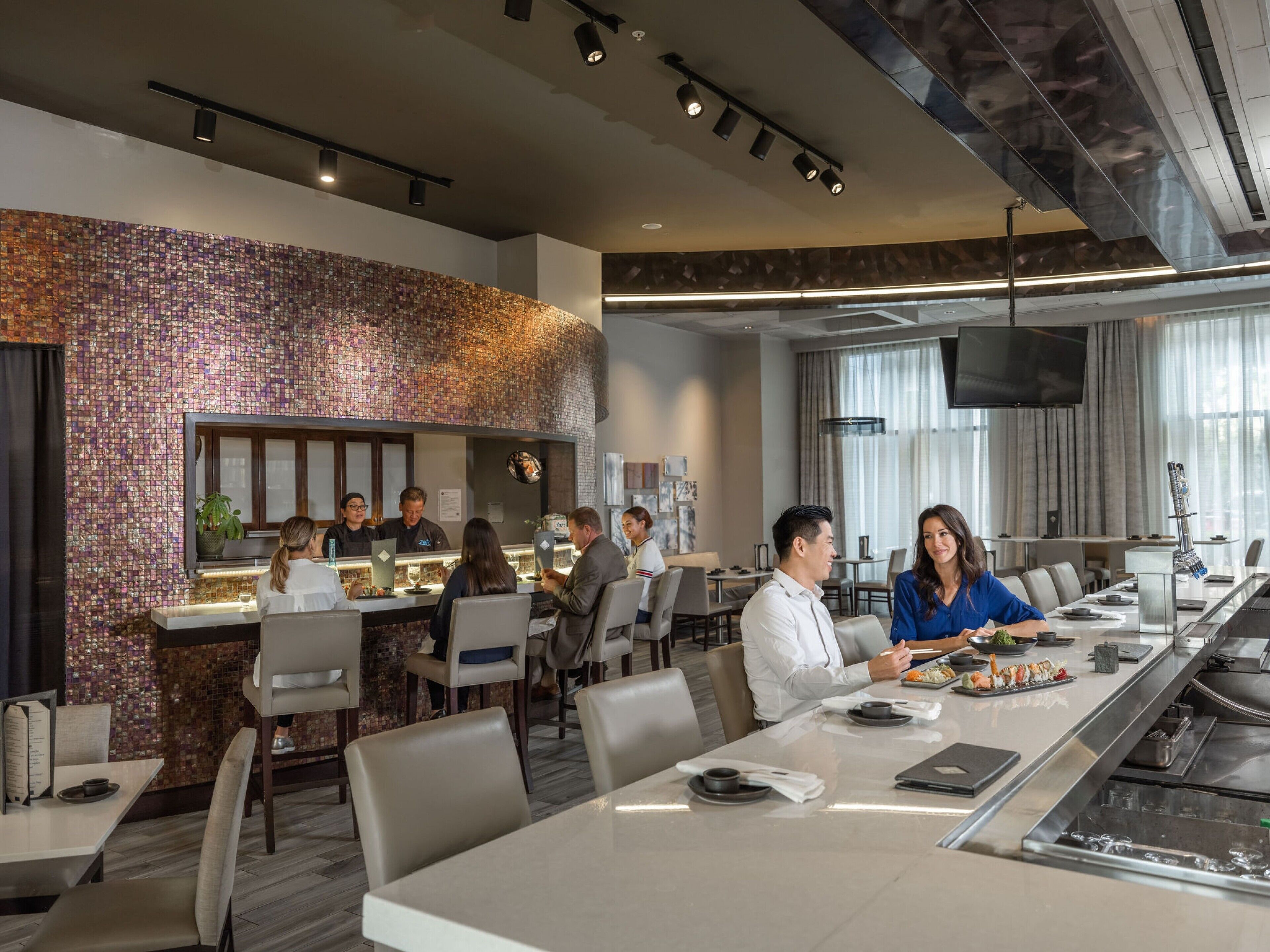 A modern restaurant with a long communal table, individual seating, a bar area, and a TV screen.