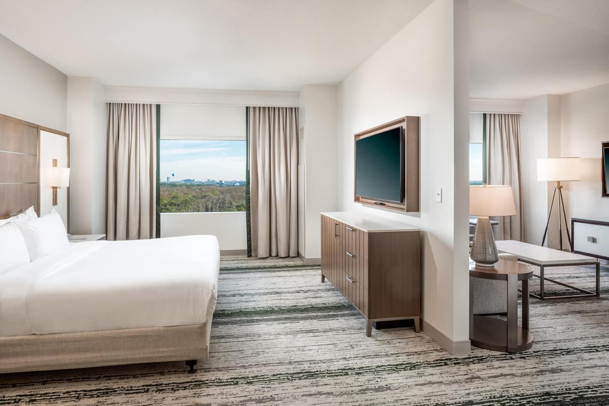Ein Hotelzimmer mit einem großen Bett, einem an der Wand montierten Fernseher, einem Schreibtisch mit Lampe und Blick auf die Stadt.